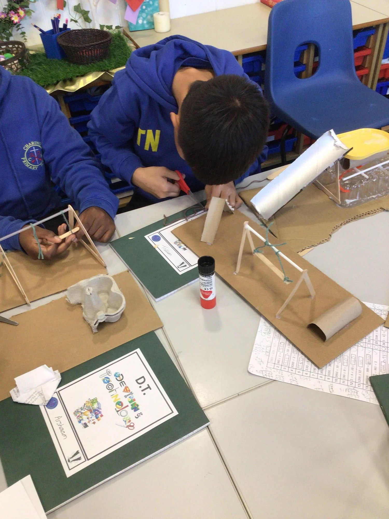 Students working on a science project with cardboard, paper towel tubes, and sticks in a classroom.