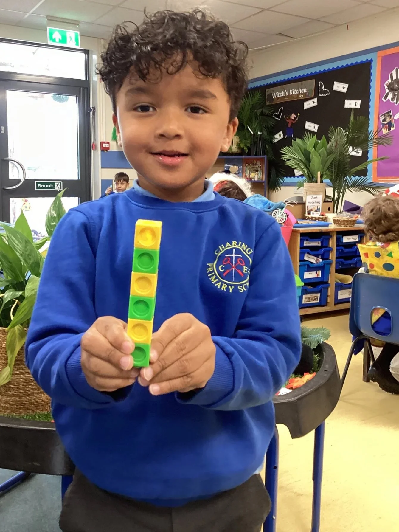 A young boy in a blue school uniform holding a colorful stacking toy in a classroom.