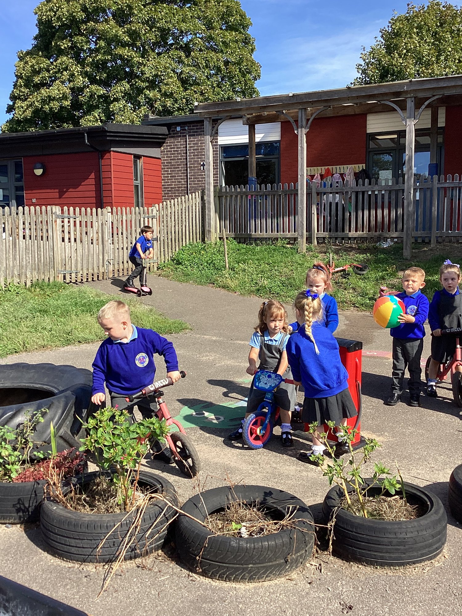 Children playing outdoors at a daycare or preschool, some on bikes and a beach ball, with tires and plants in the foreground and a wooden fence and building in the background.