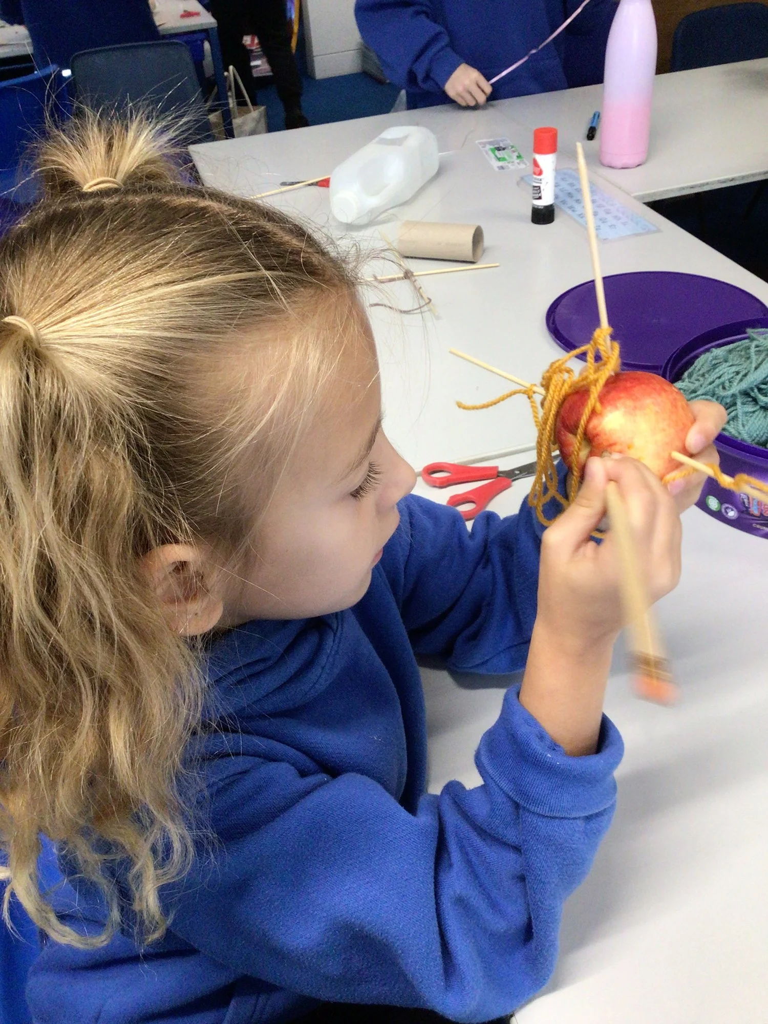 A girl with blonde, curly hair and in Charing Primary School uniform is sitting at a table, wrapping yarn around an apple with a wooden skewer. There are scissors, glue, and craft supplies on the table.