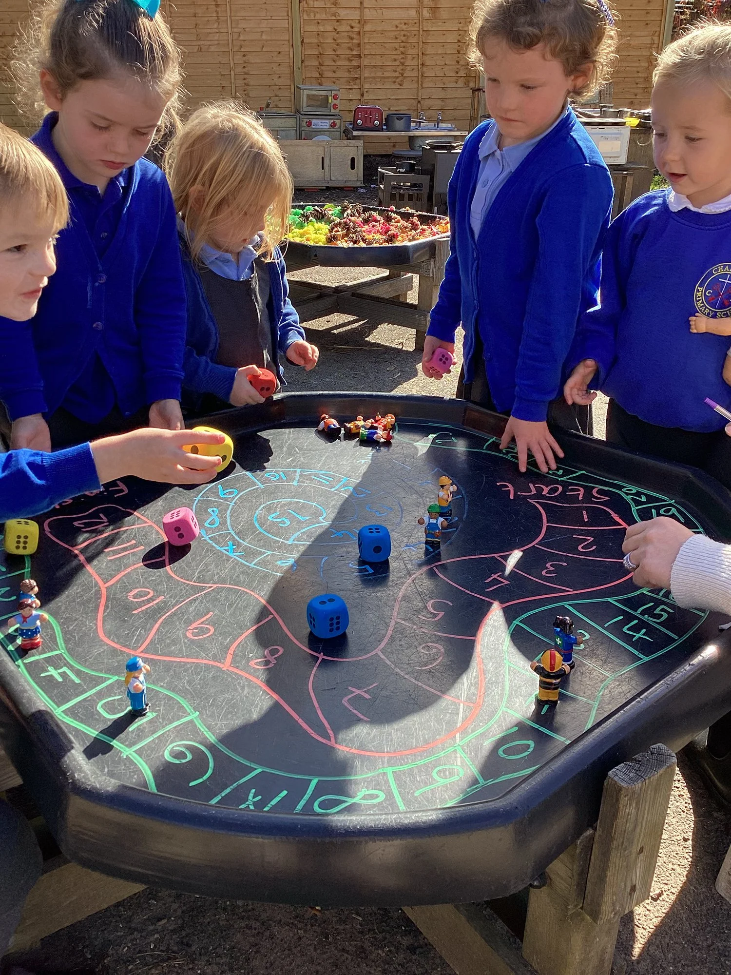 Children playing a dice game on an outdoor table, surrounded by toys and colorful figures, with a wooden fence in the background.