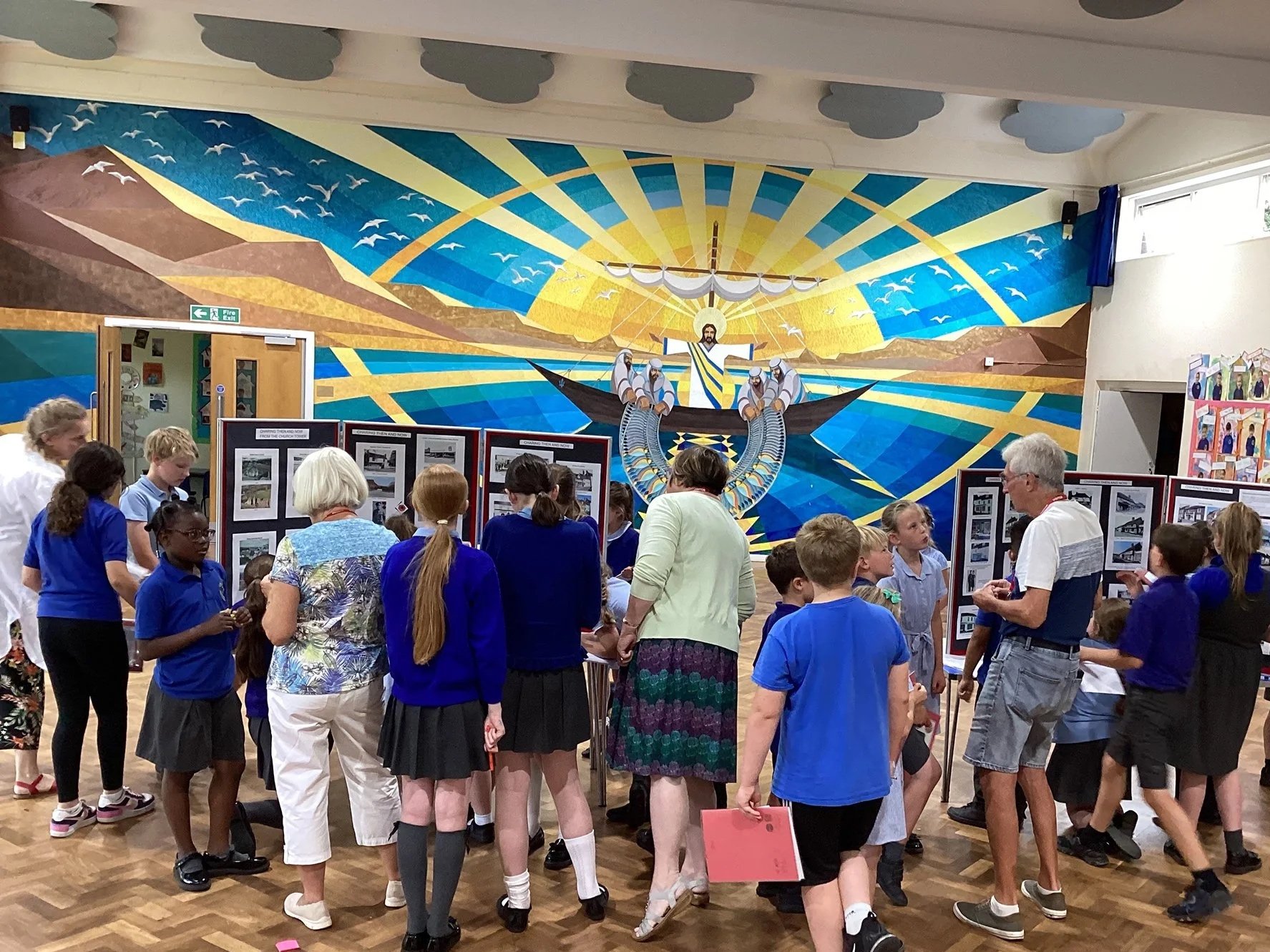 A group of children and adults gathered in a classroom or gallery, looking at display boards with photographs or artwork. The background features a large mural of a religious scene with a boat and a radiant sun.
