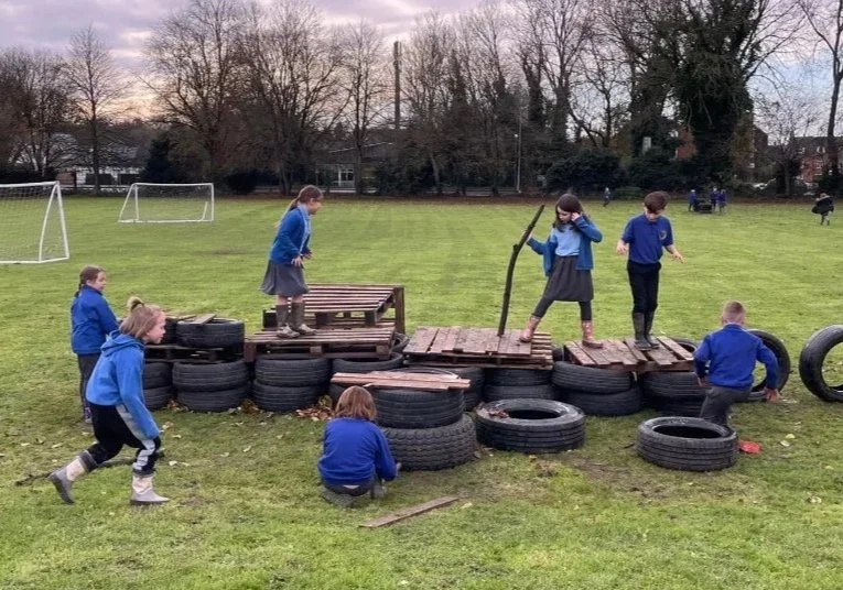 School children playing on a makeshift fort built with tires, wooden pallets, and a stick in a grassy field.
