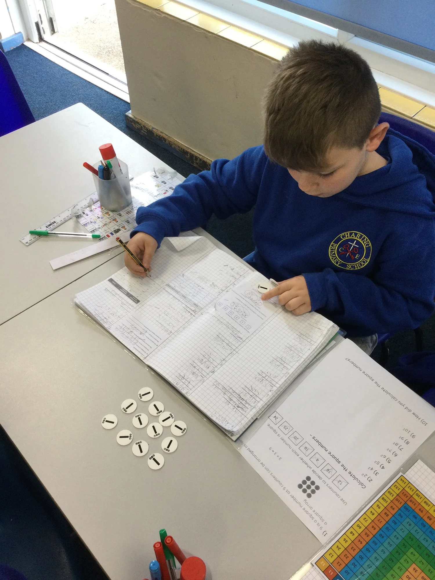 A boy in a blue school uniform sitting at a desk, working on math homework with graph paper, with counting tokens, pens, ruler, and educational charts on the desk.