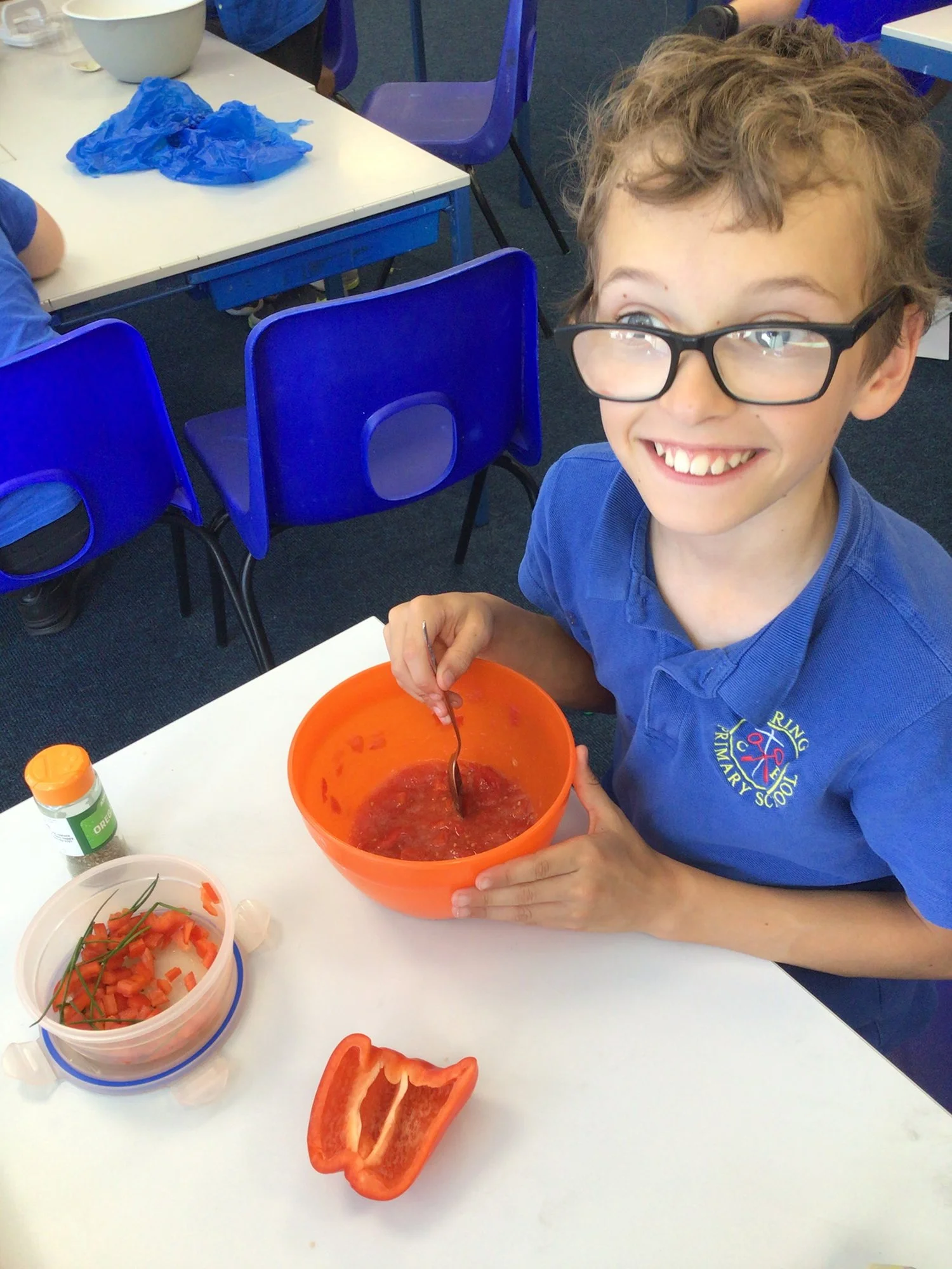 A young boy with glasses smiling and stirring a bowl of red jelly at a classroom table, with chopped red bell pepper and a small container of chopped vegetables on the table.