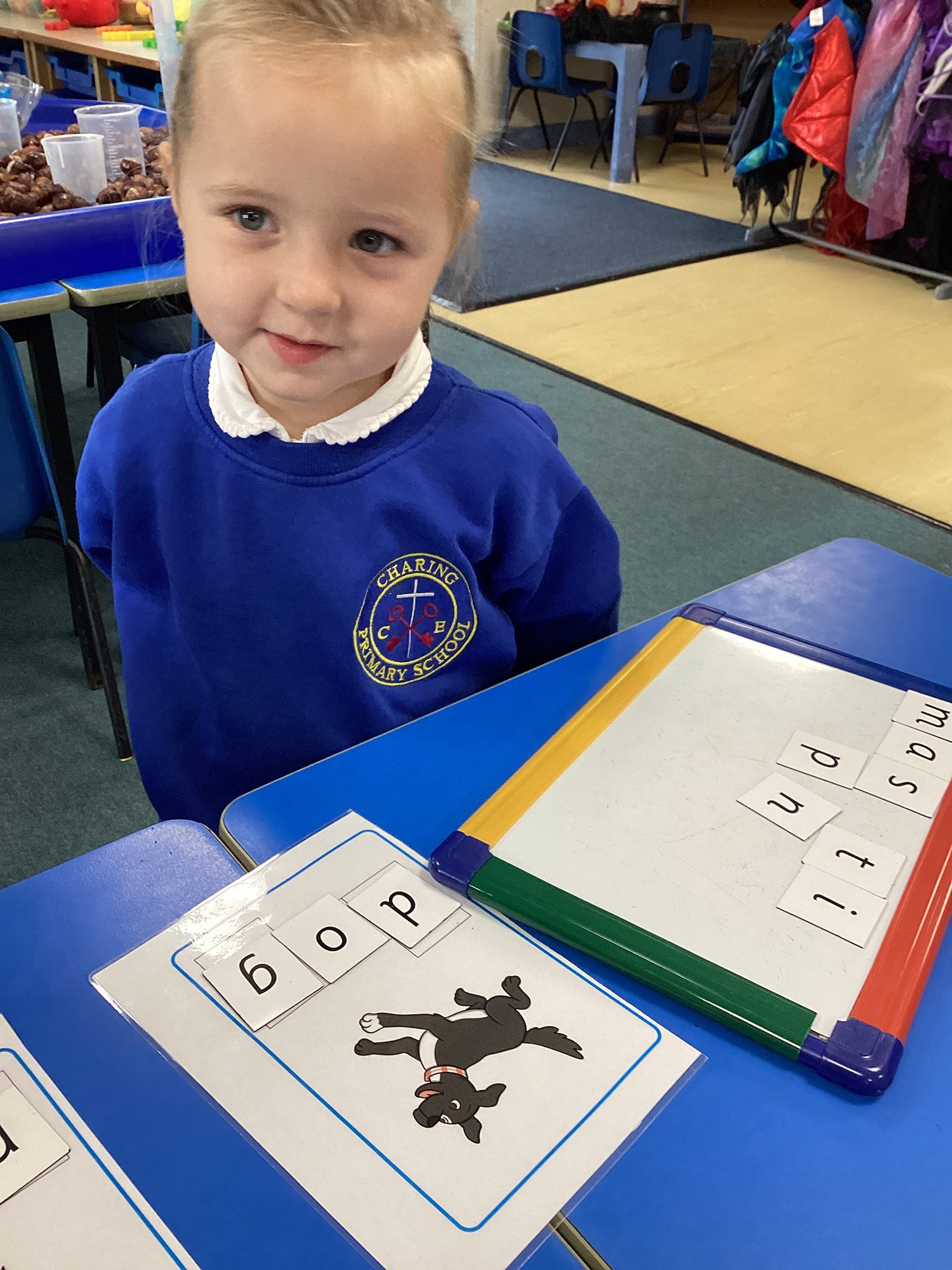 A young girl in a blue school uniform sitting at a desk with a laminated card featuring the word "bop" and a picture of a black dog. There are magnetic letter tiles on a dry erase board.