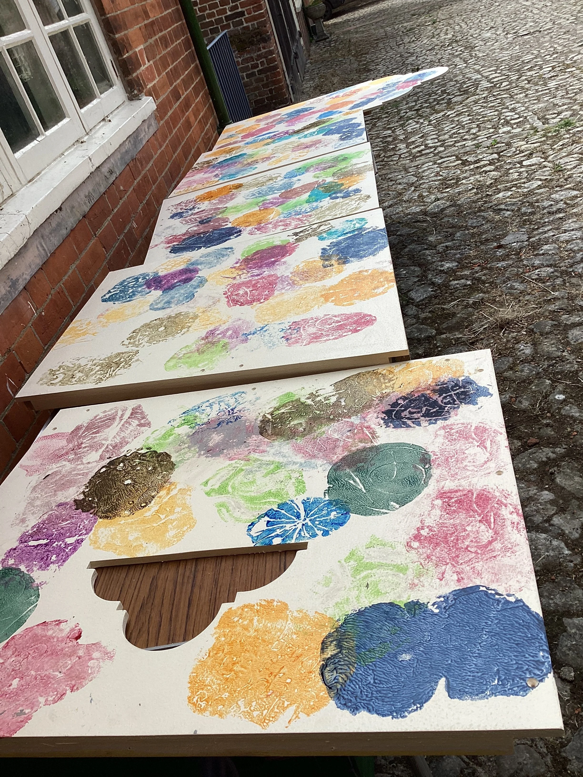 Outdoor artwork on wooden panels featuring colorful abstract prints, including circular and textured patterns, laid on a cobblestone ground next to a brick building.