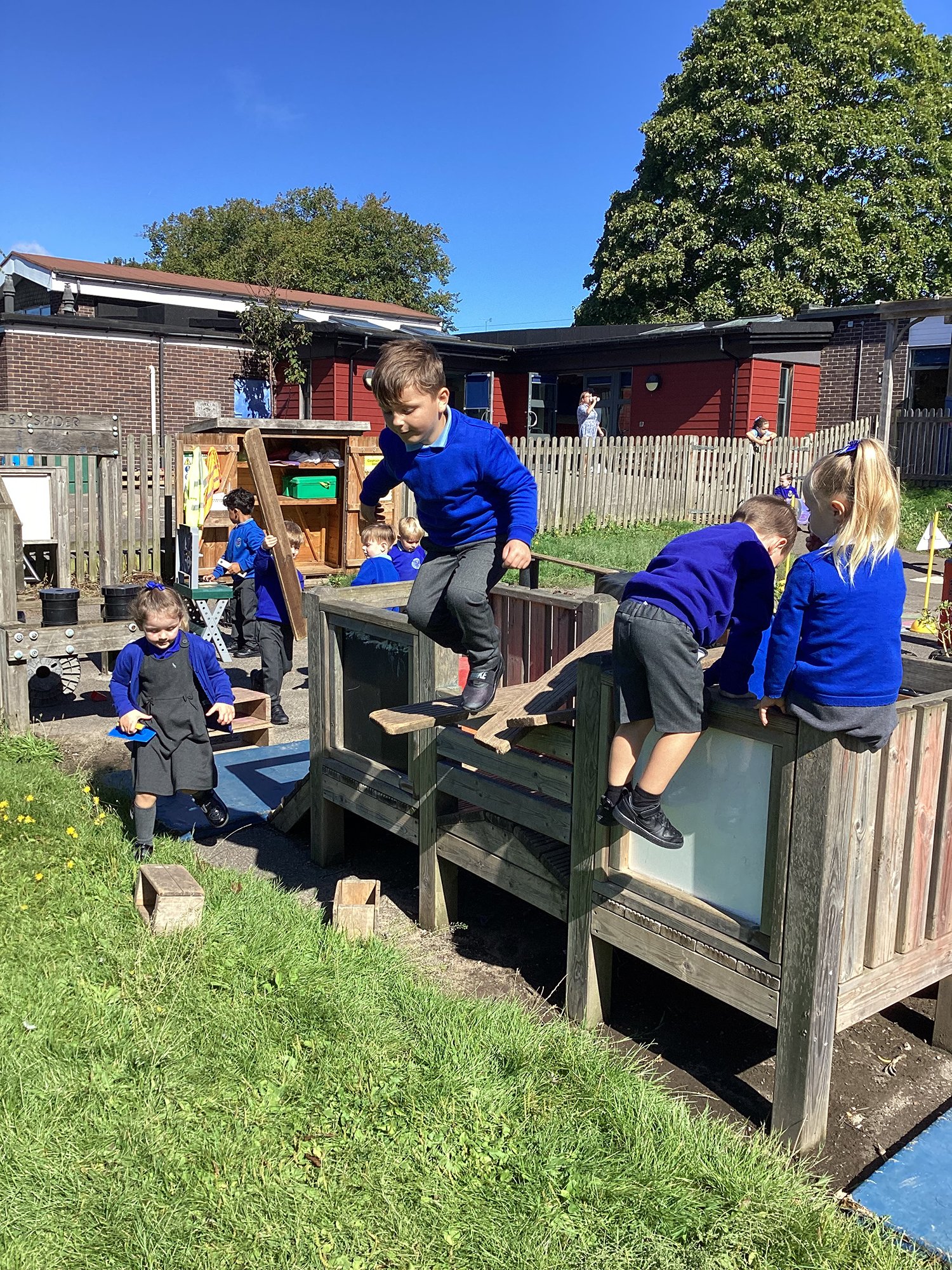 Children playing outside on a wooden playground structure at a school or park with trees and buildings in the background, on a sunny day.