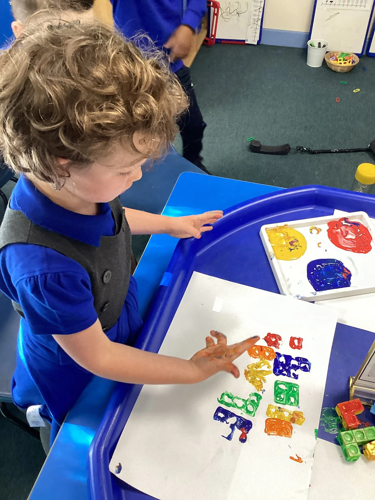 A young child with curly hair, wearing a blue uniform and gray vest, is finger painting with colorful paints on a white sheet of paper at a blue table in a classroom.