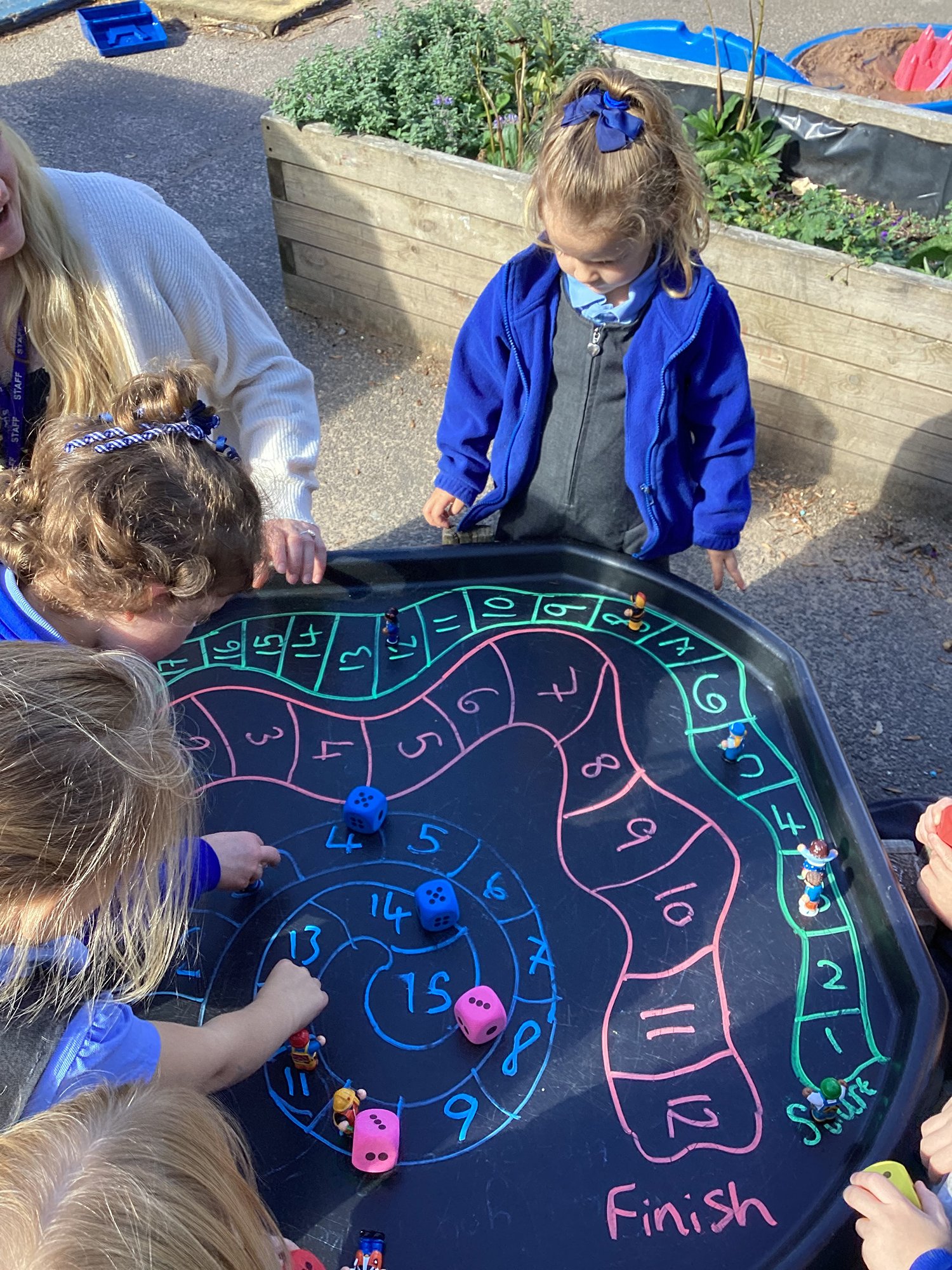 Children and an adult playing a colorful board game outdoors on a black table with a spiral track and numbered spaces, several dice, and game pieces, with greenery and garden beds in the background.
