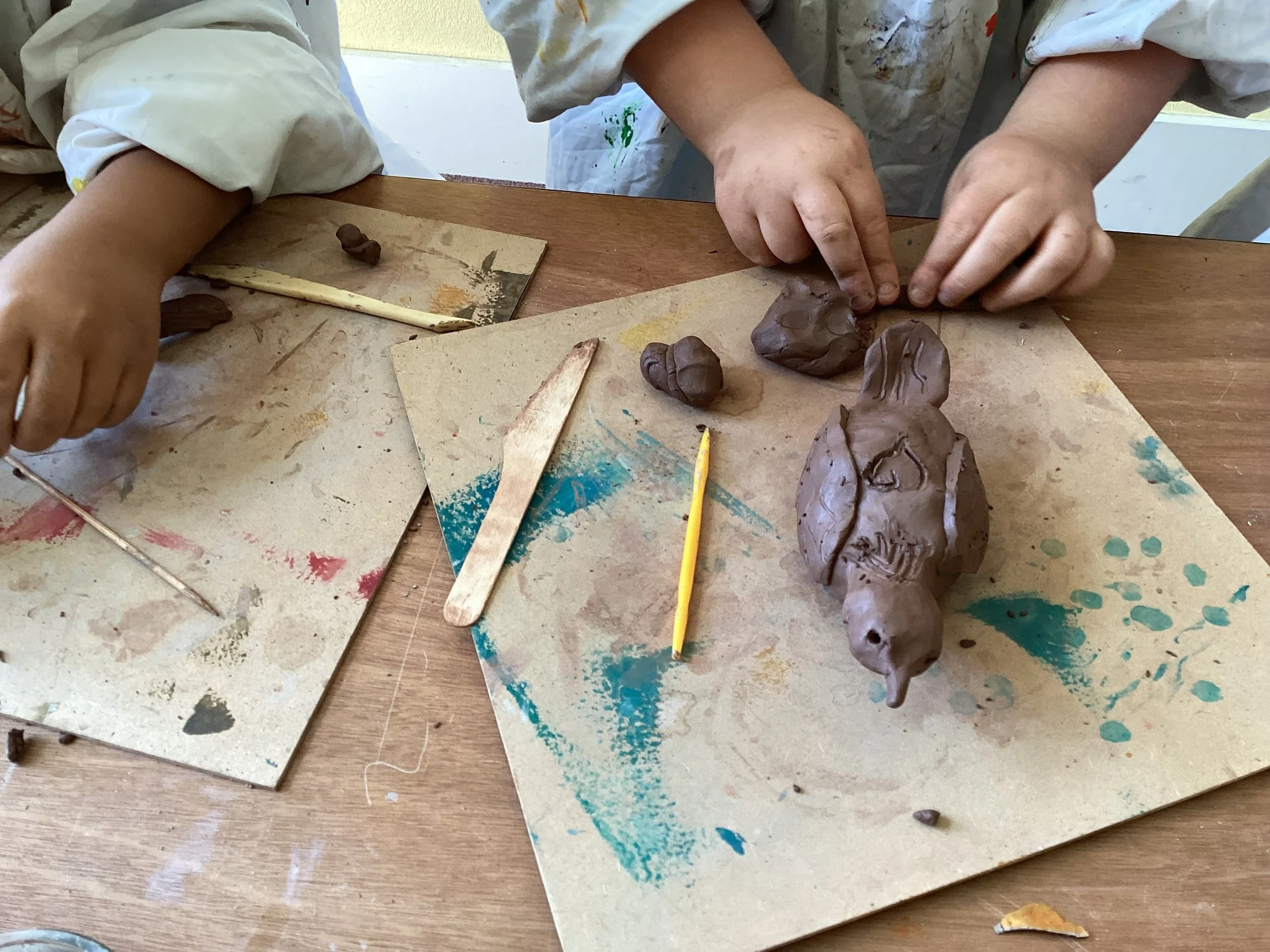 Children working on a clay sculpture project at a wooden table, with clay, sculpting tools, and protective smocks.