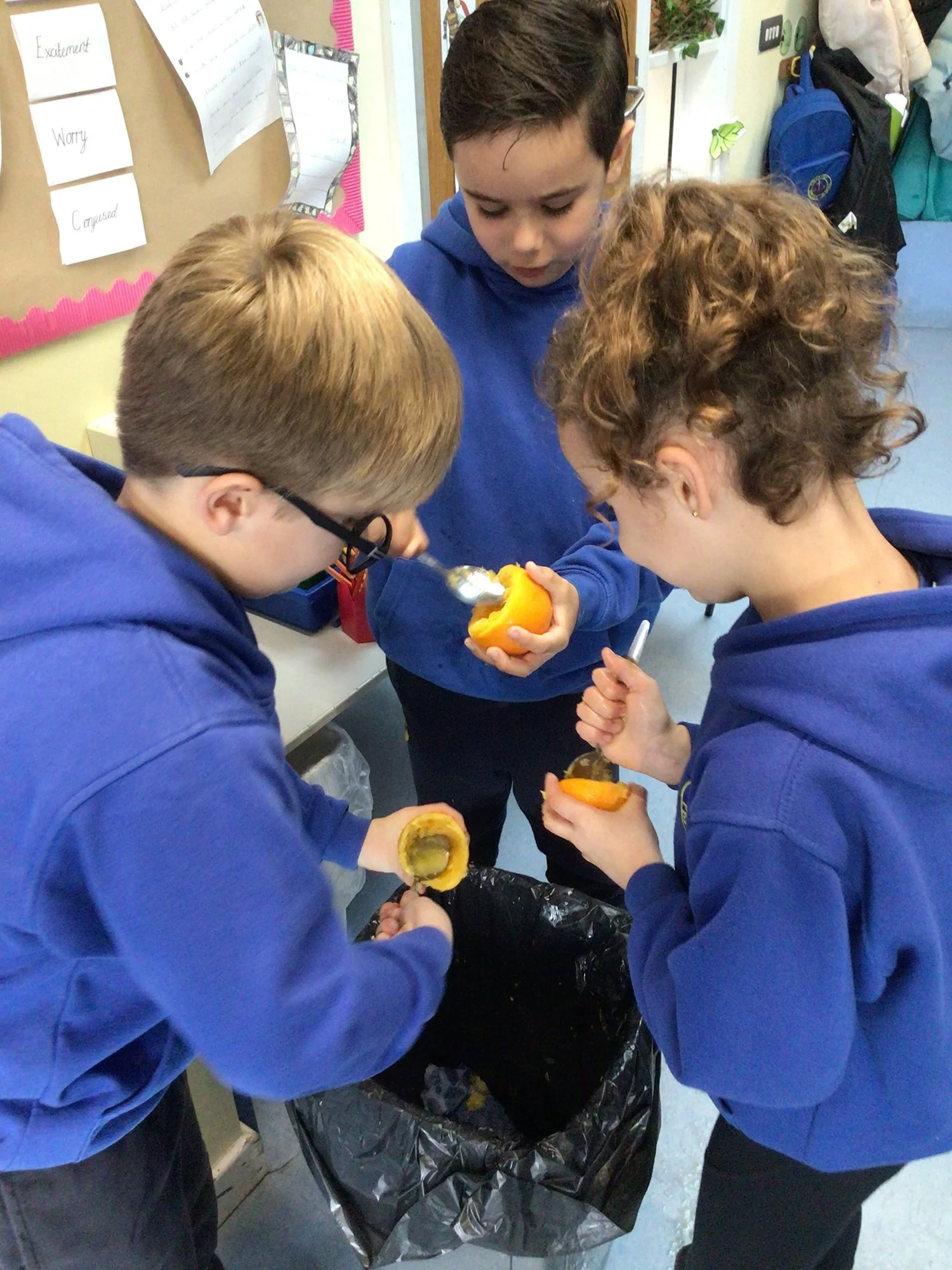 Three children wearing blue hoodies are gathered around a trash bin, peeling and scooping out the insides of orange pumpkins into the trash.
