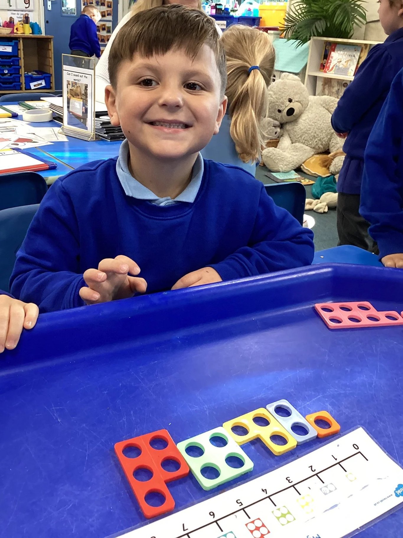 A young boy sitting at a classroom table smiling, with colorful counting blocks and a number chart in front of him.