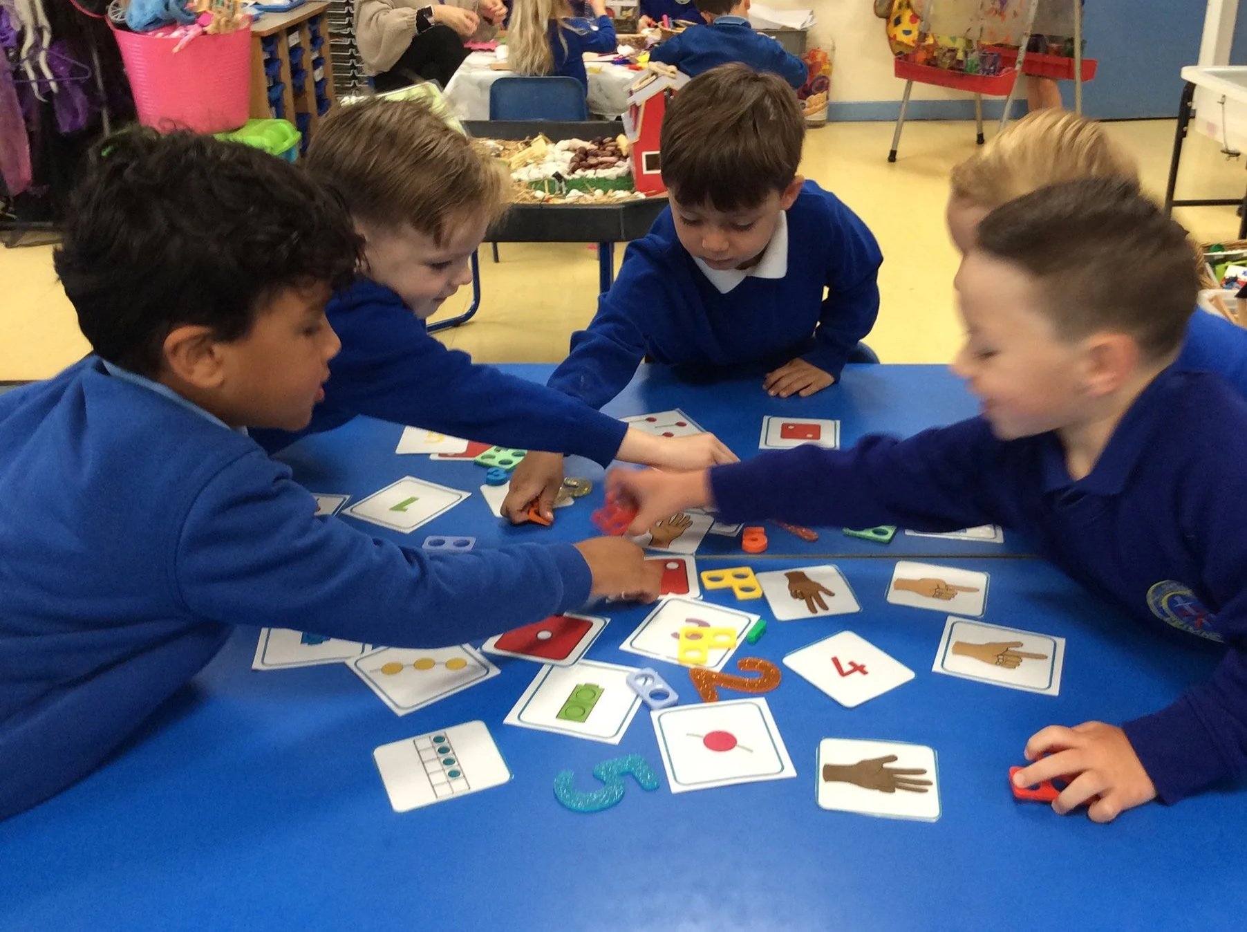 Five children in blue uniforms are playing a card game on a blue table in a classroom. They are reaching for cards and appear engaged in the game. The background shows other children, tables, and classroom supplies.