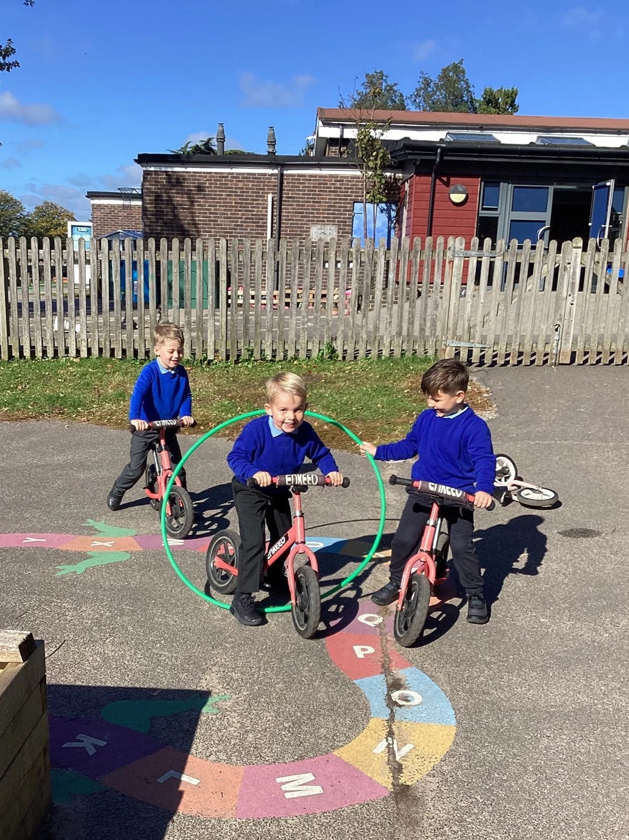 Three young boys wearing blue sweaters playing outside on scooters and a hula hoop on a colorful hopscotch pattern on the ground.