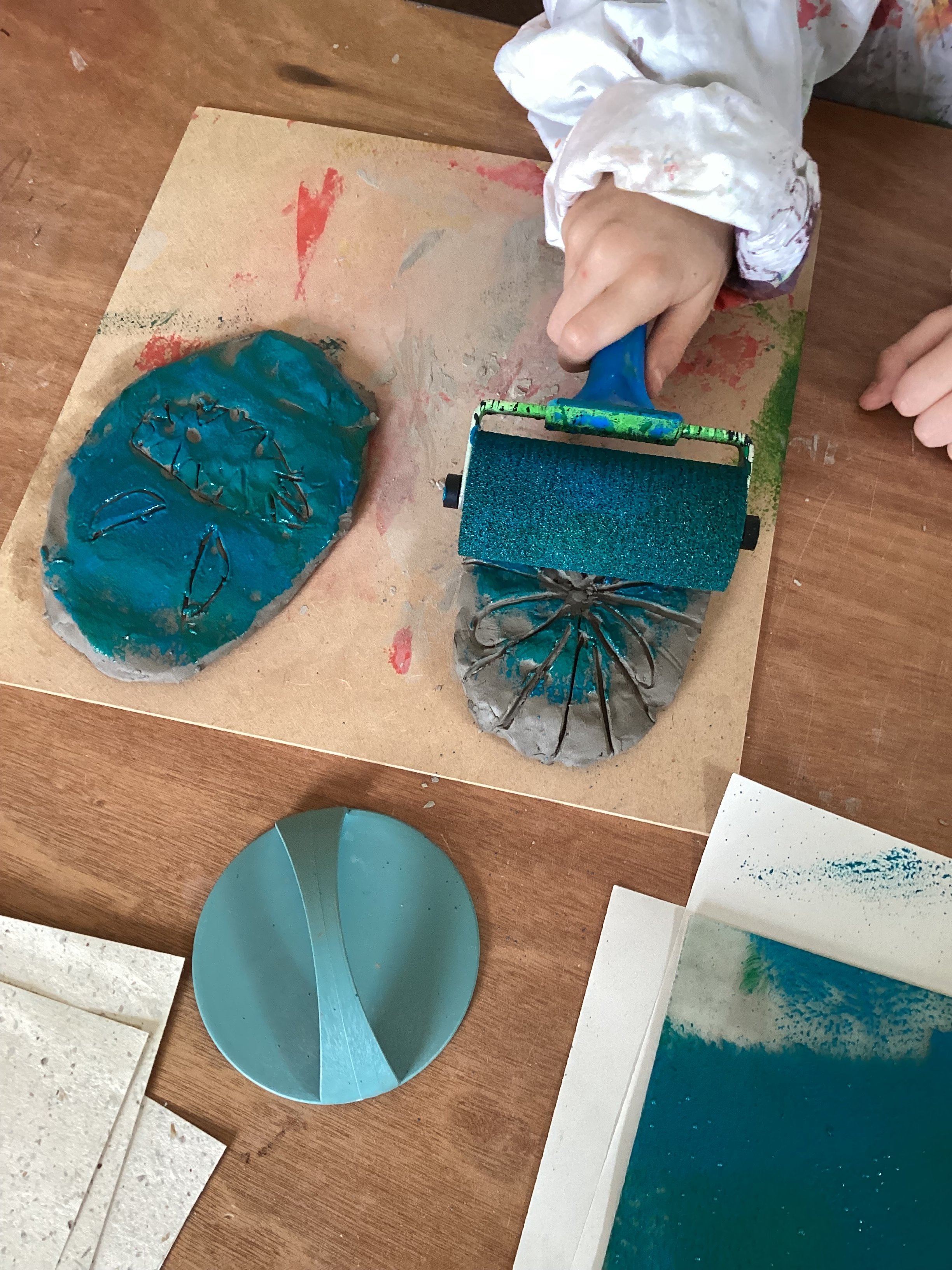Child using a brayer to print a butterfly design on a rock with turquoise paint, on a wooden table.