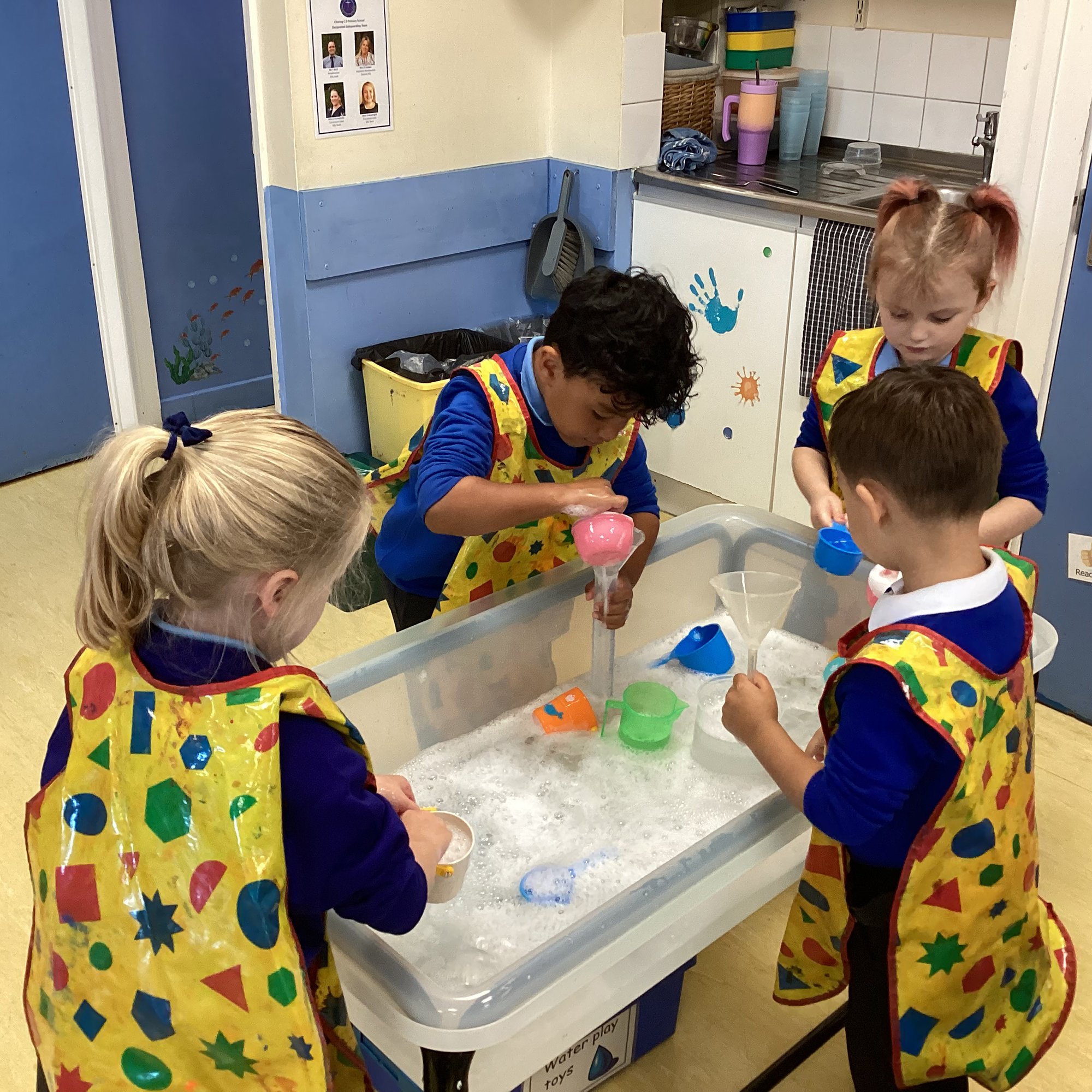 Four young children wearing colorful aprons are playing with water and toy cups at a water play table in a classroom.