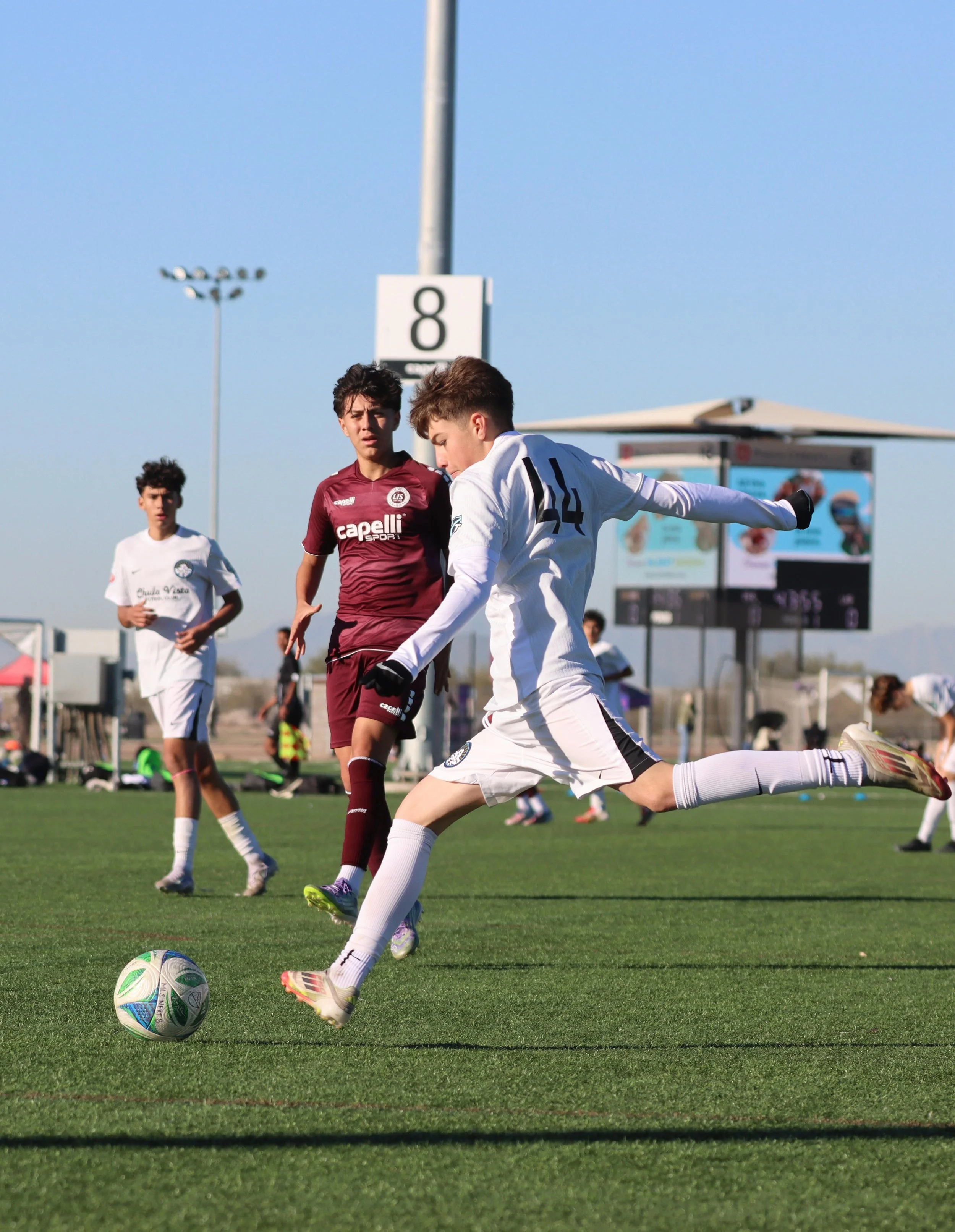 Soccer players in white and maroon jerseys competing for the ball on a green field, with digital scoreboards and advertisements in the background under a clear sky.