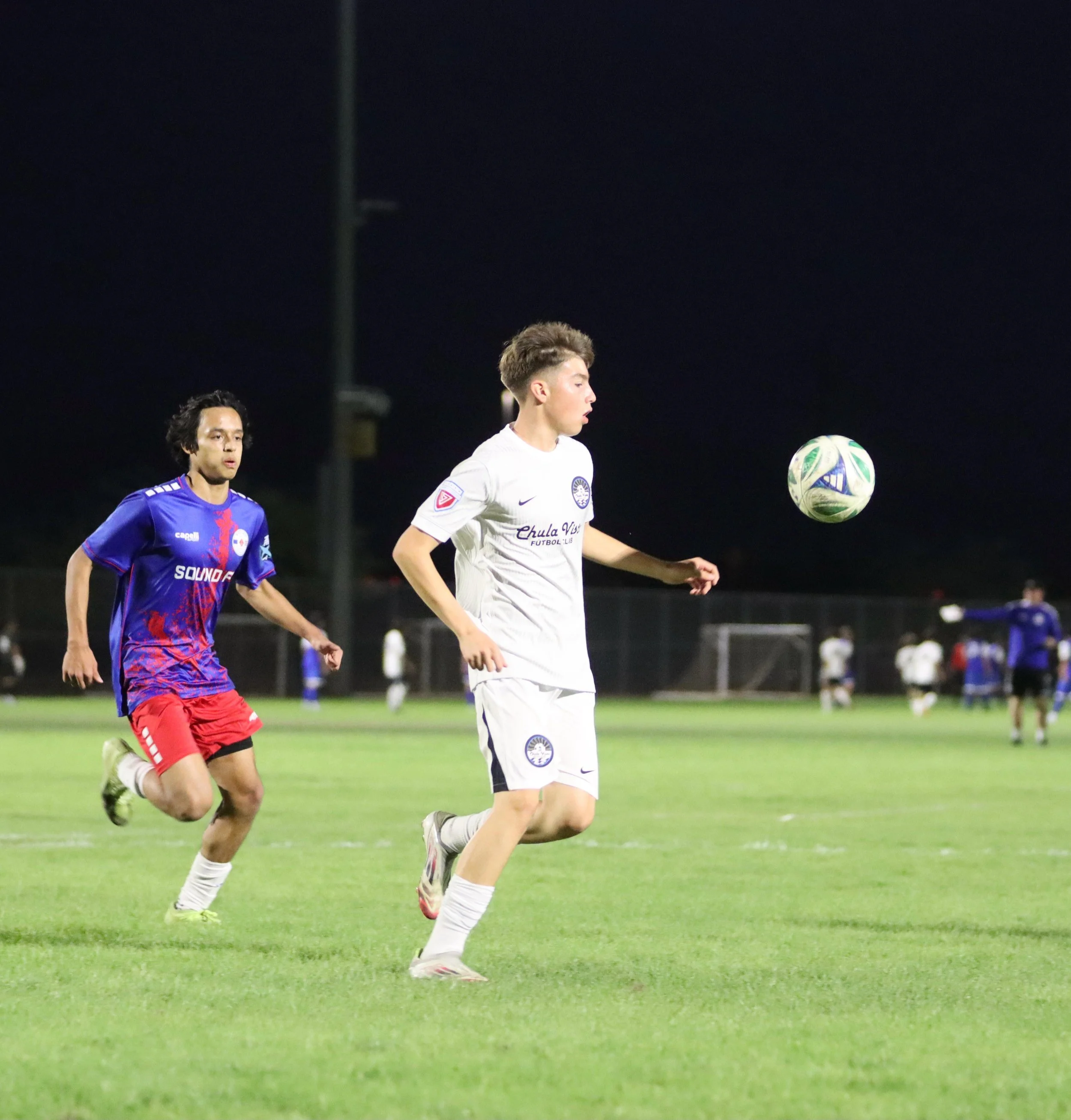 Two soccer players chasing a ball on a green field at night, with other players visible in the background.