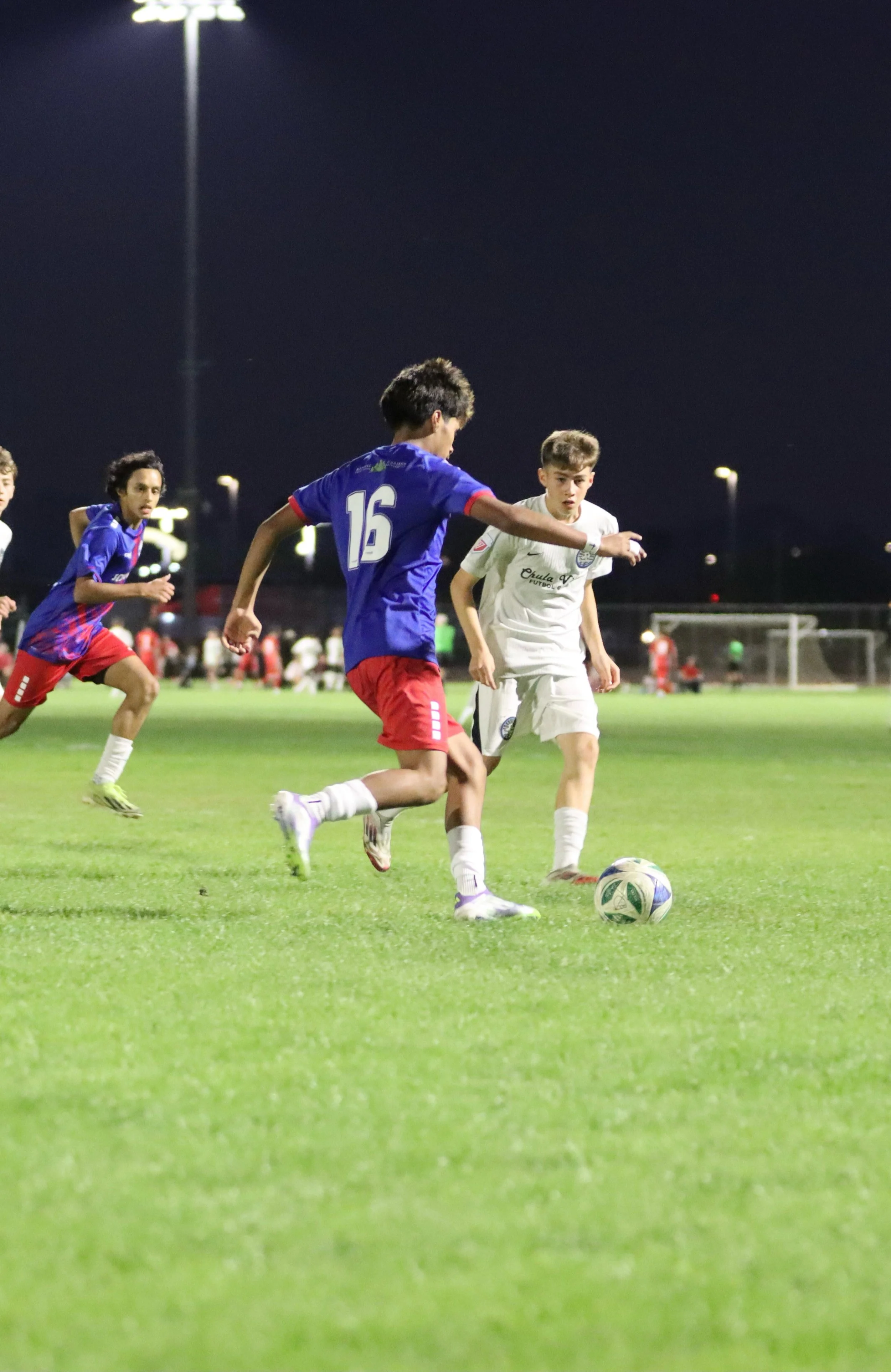 Young soccer players competing on a well-lit field at night, with two boys focused on kicking the ball while others run nearby.