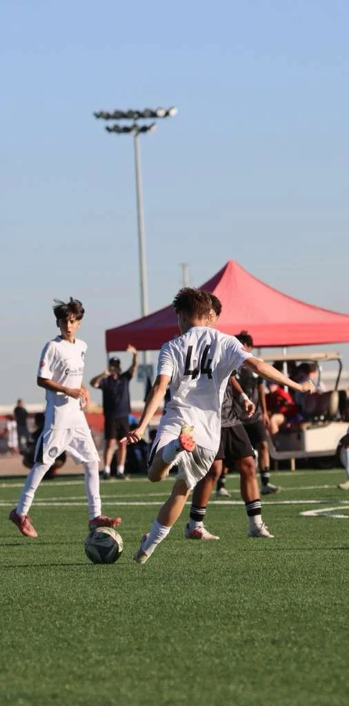 Young soccer players in white uniforms playing on a green field during daytime with a red tent and other people in the background.
