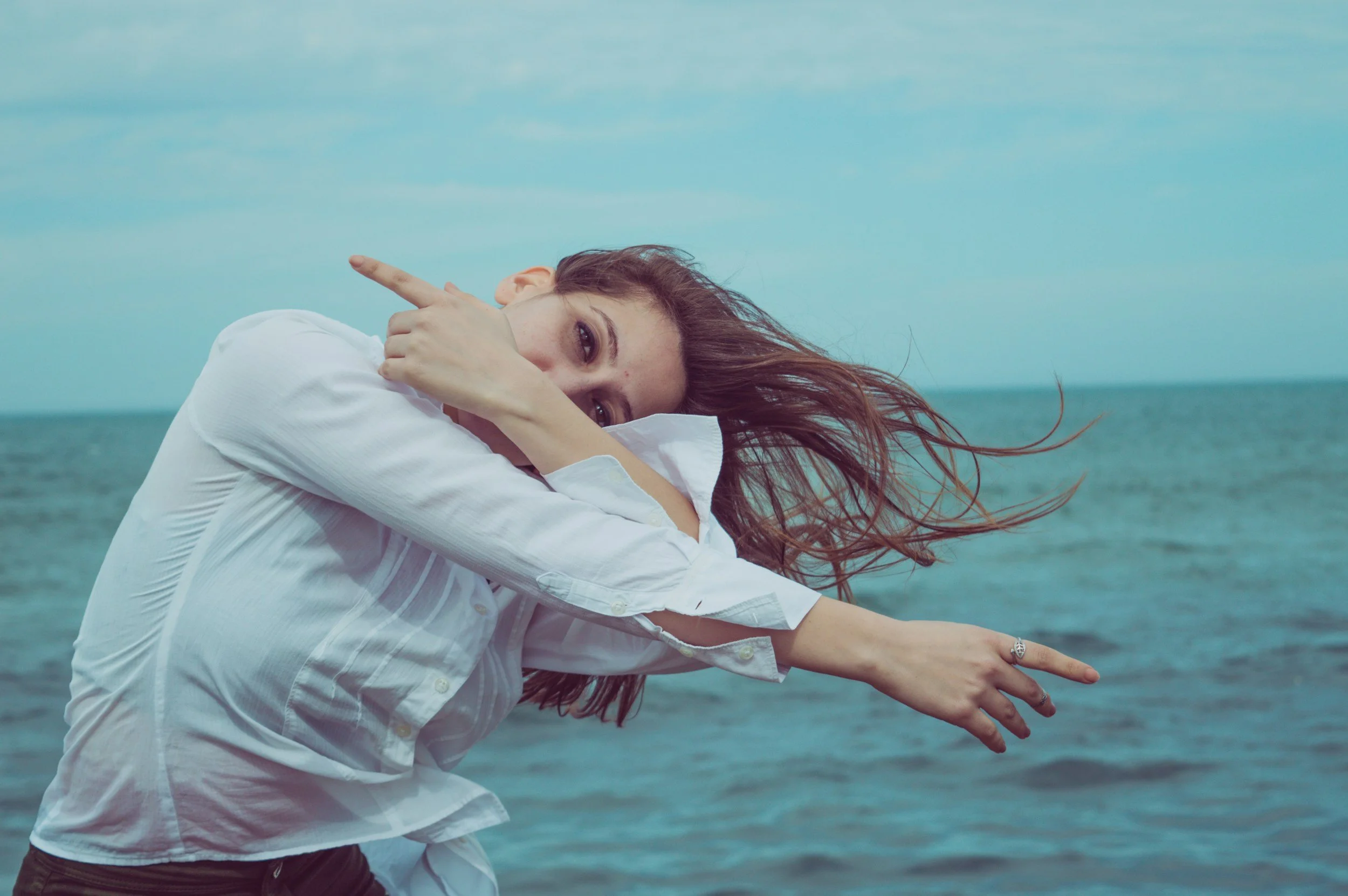 A woman with red hair wearing a white shirt is leaning forward on a dock by the water, with her arm extended in two different directions, and the sea and sky in the background.