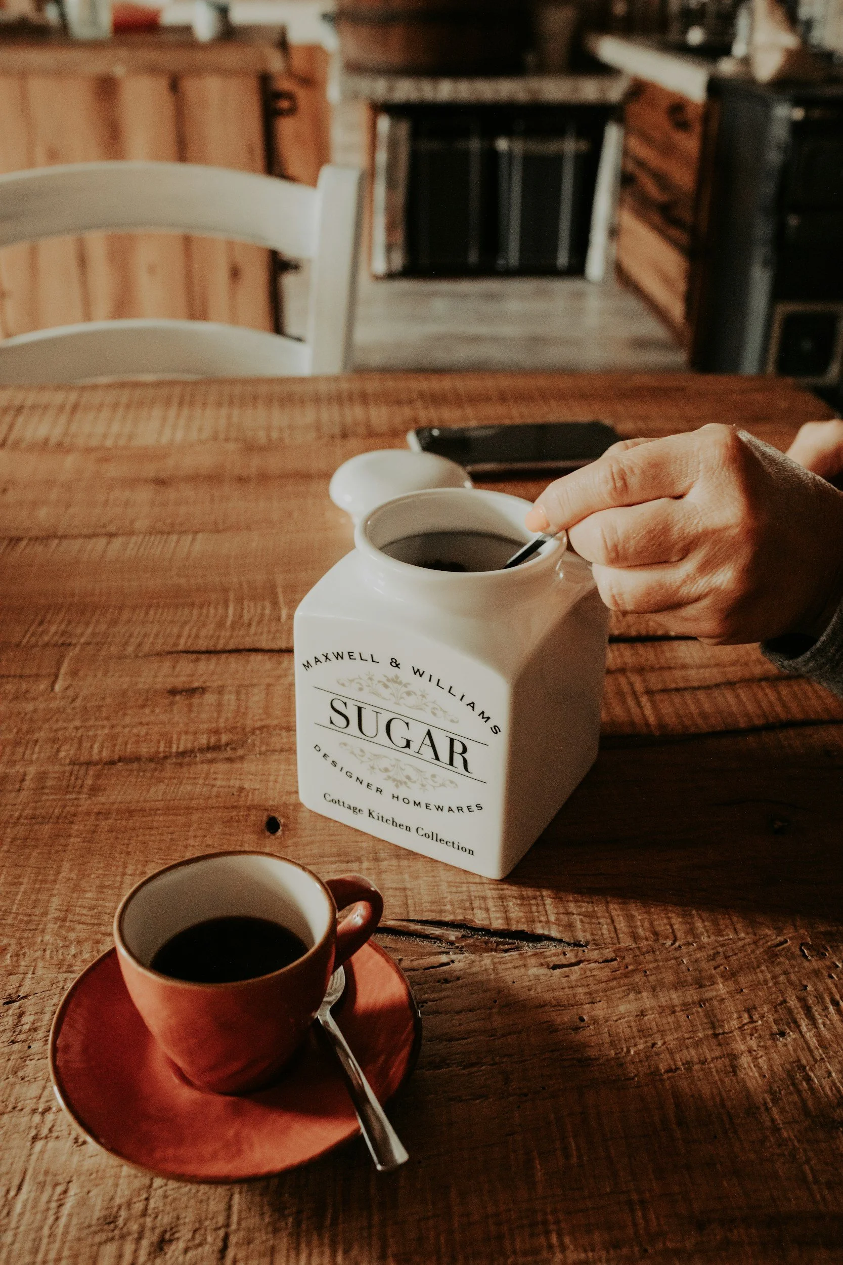 A wooden table with a white ceramic sugar container, a small brown cup, and a person's hand holding a spoon over the sugar container, with a background of kitchen cabinets and a stove.