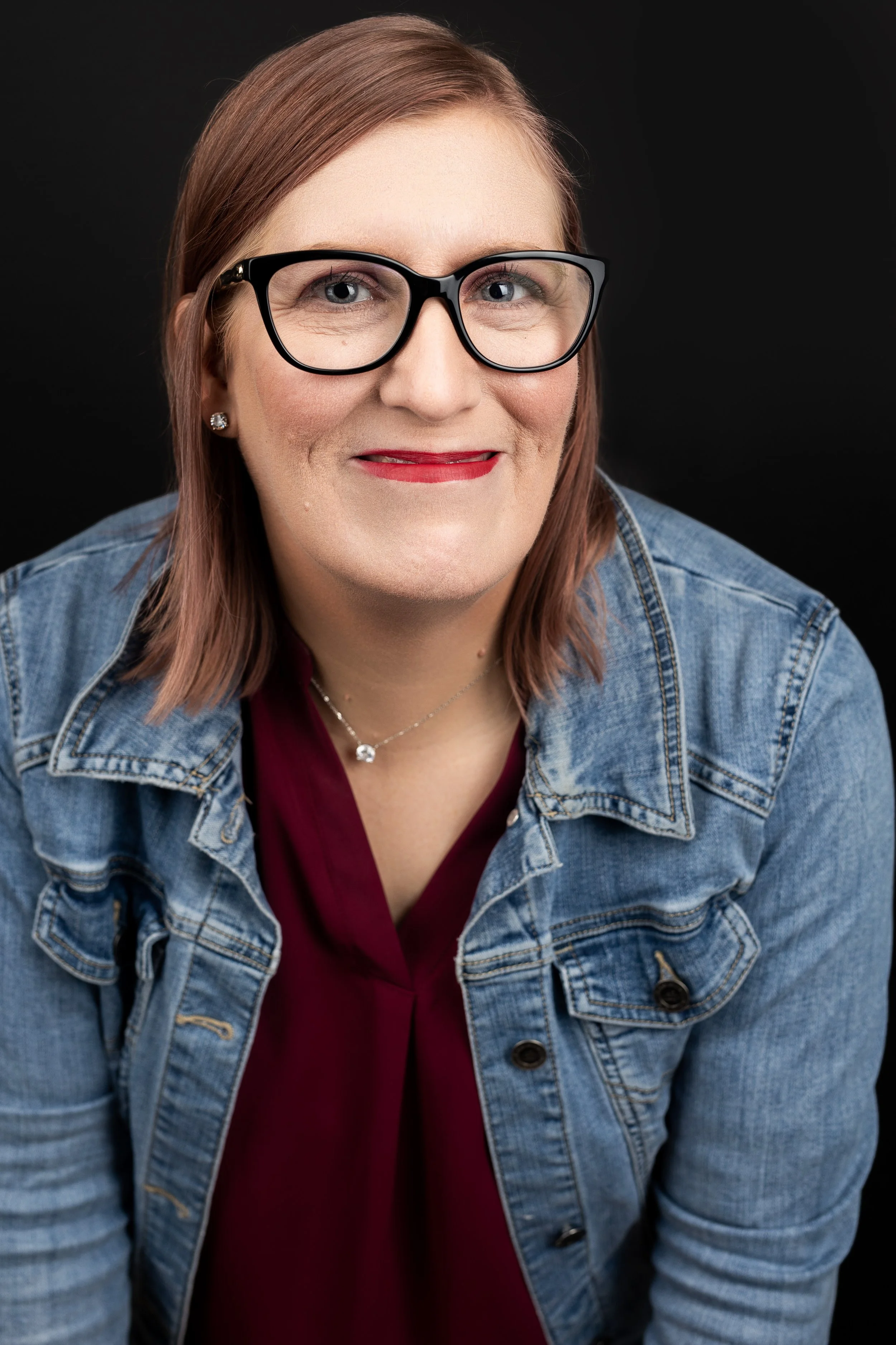 A woman with reddish hair, wearing glasses, a denim jacket, red lipstick, and a burgundy top, smiling against a black background.