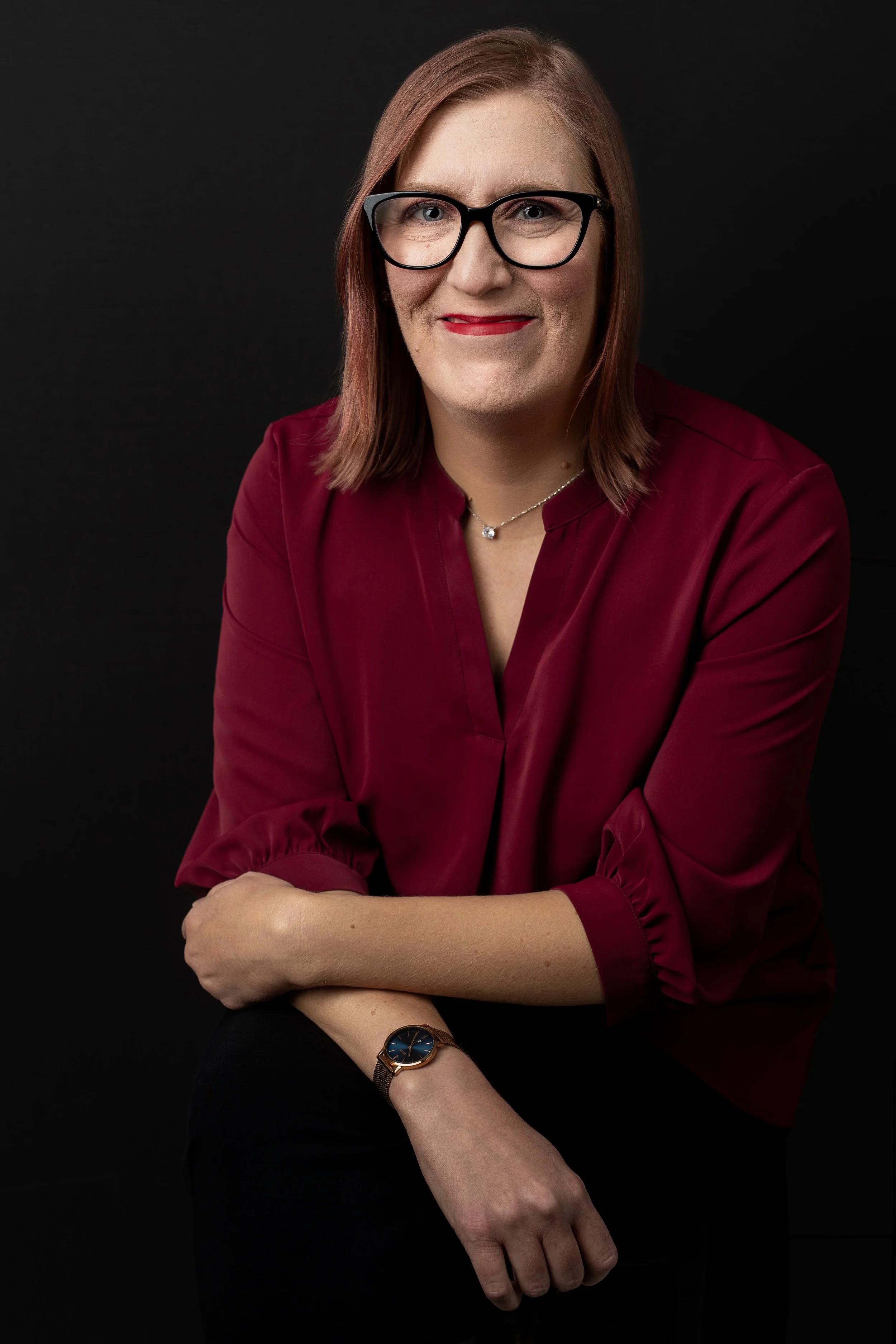 Portrait of a woman with shoulder-length brown hair, wearing black glasses, a red blouse, a silver necklace with a small pendant, and a watch with a blue face, posing against a black background.