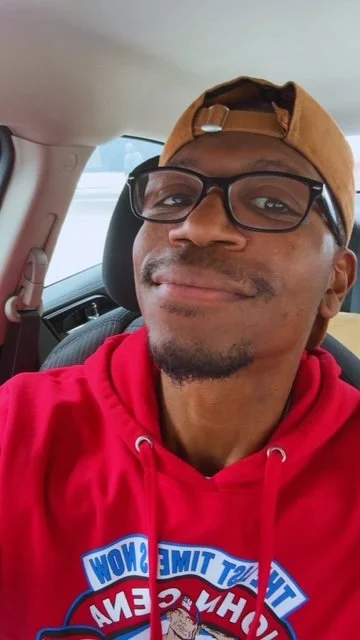 A man wearing glasses, a brown baseball cap backwards, and a red hoodie sitting in a car, smiling at the camera.