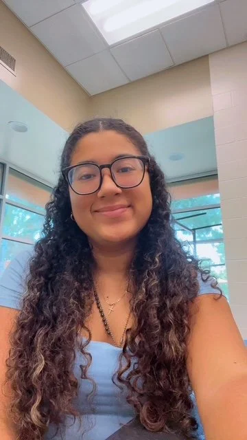 Young woman with long curly hair and glasses smiling in an indoor setting with large windows and a beige and light blue wall.