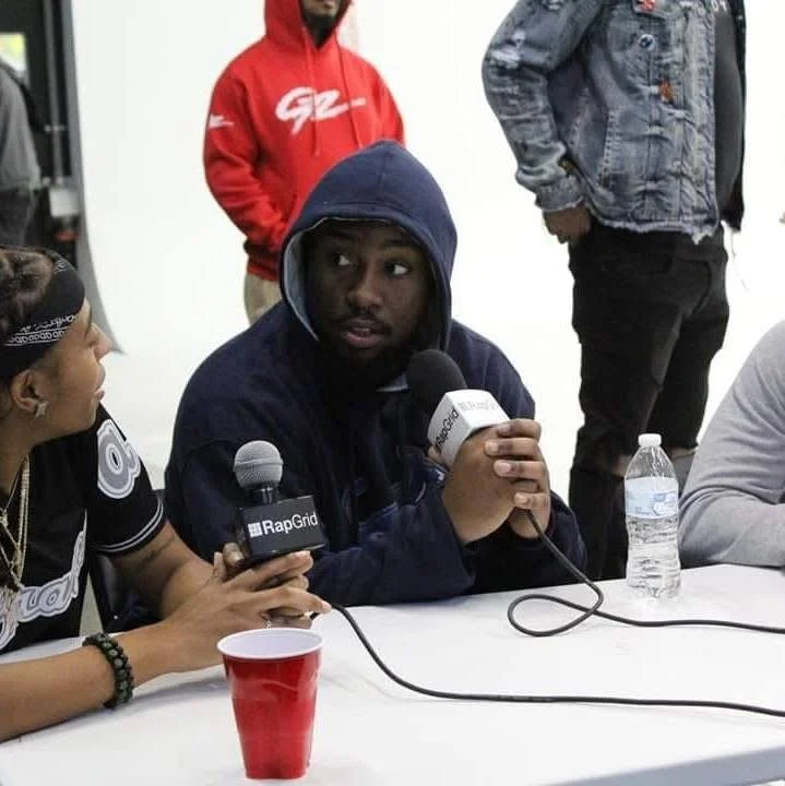 A young man in a dark hoodie speaks into a microphone during an interview. He is sitting at a table with another person holding a microphone, with a red plastic cup and a water bottle on the table. Several people are standing in the background.