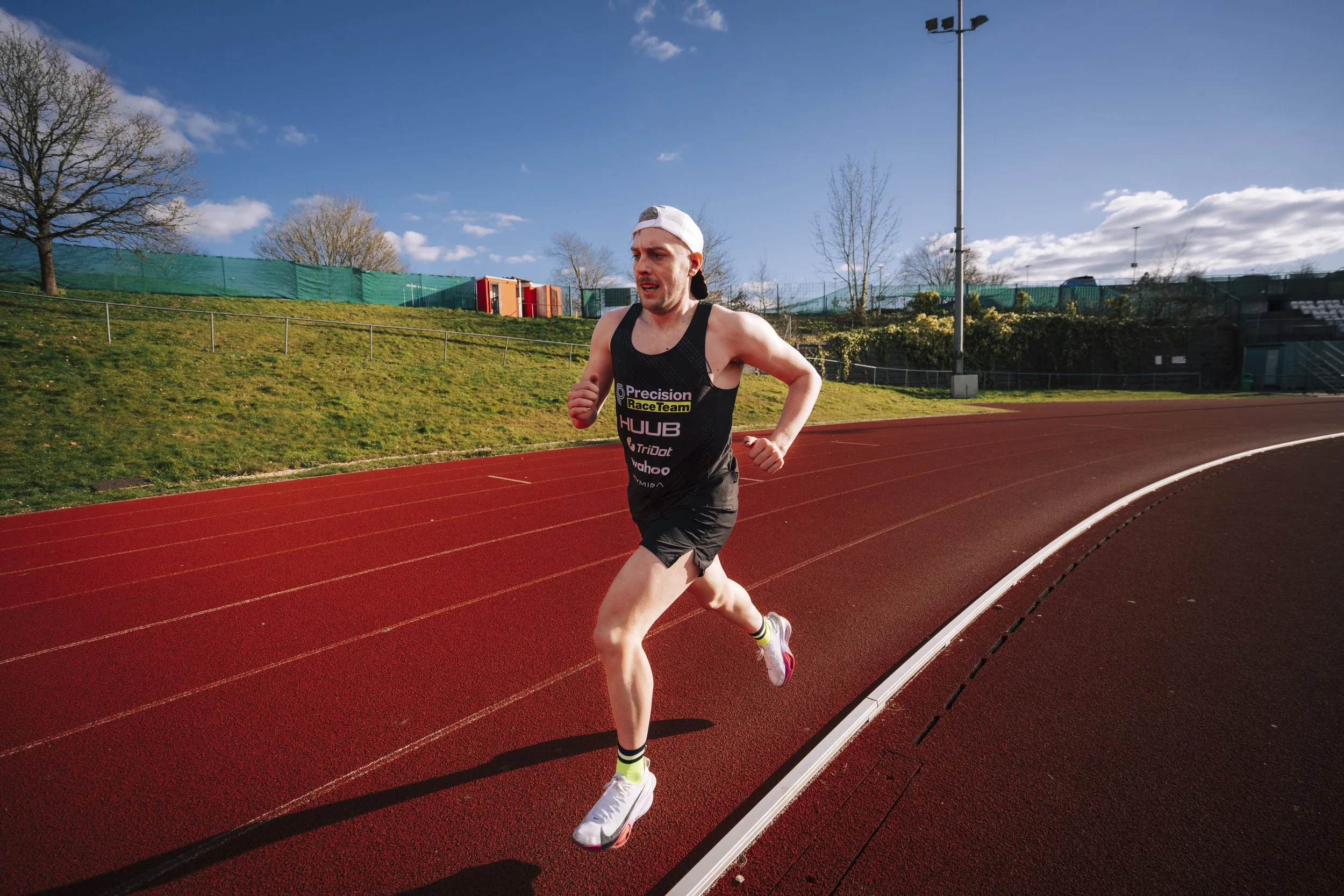 Male runner in black tank top and shorts running on an outdoor track with green grass and trees in the background under a clear blue sky.