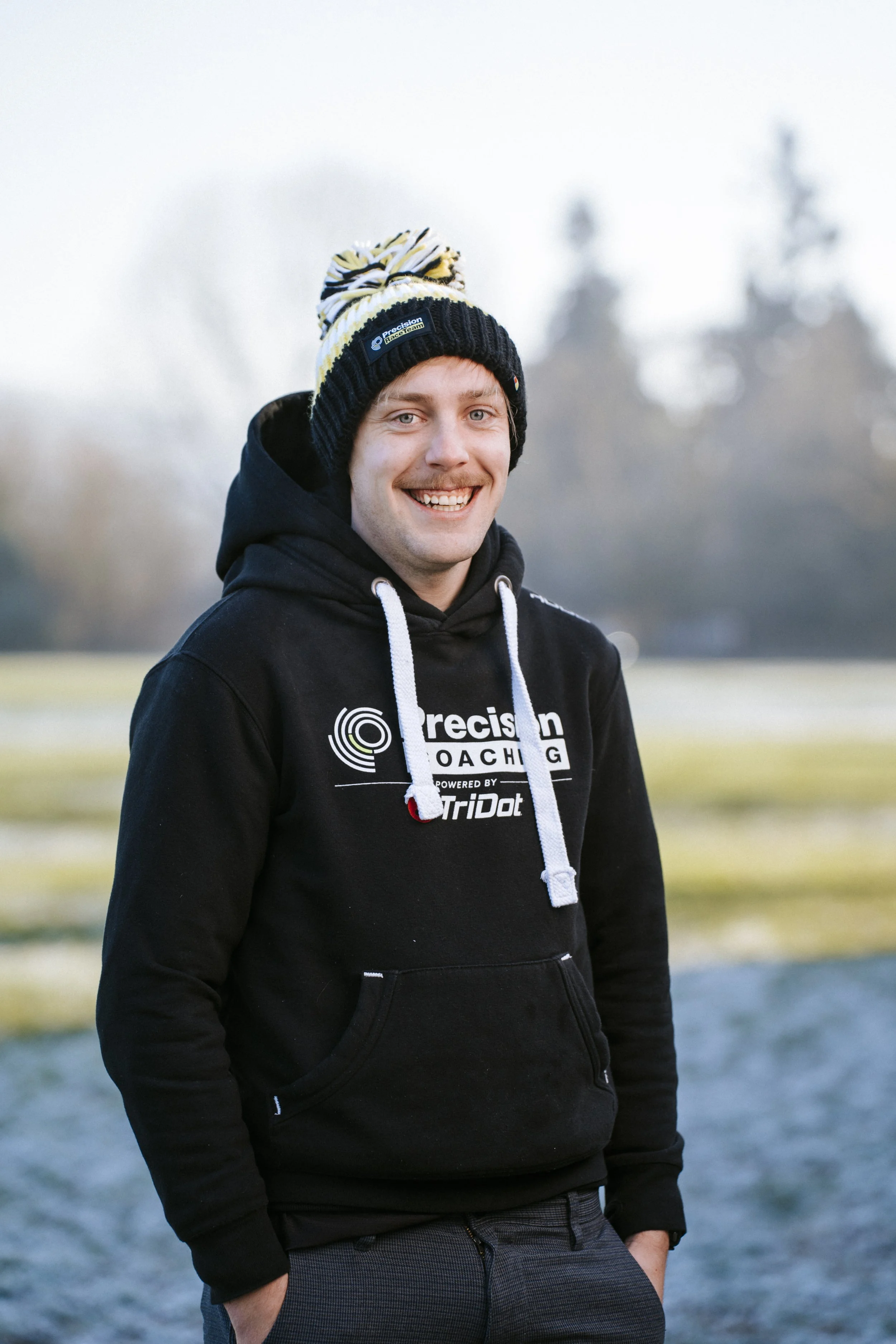A smiling young man wearing a black hoodie with white text and logos, a black winter hat with yellow, white, and black stripes, outdoors during daytime, with a blurred background of trees and sky.