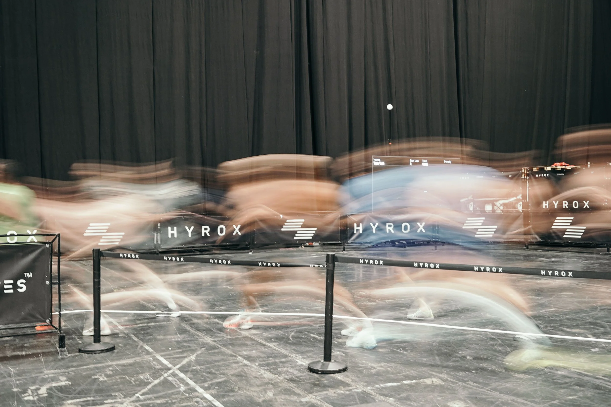 Blurred motion of people running in an indoor track with black curtains and branding that reads 'HYROX'.