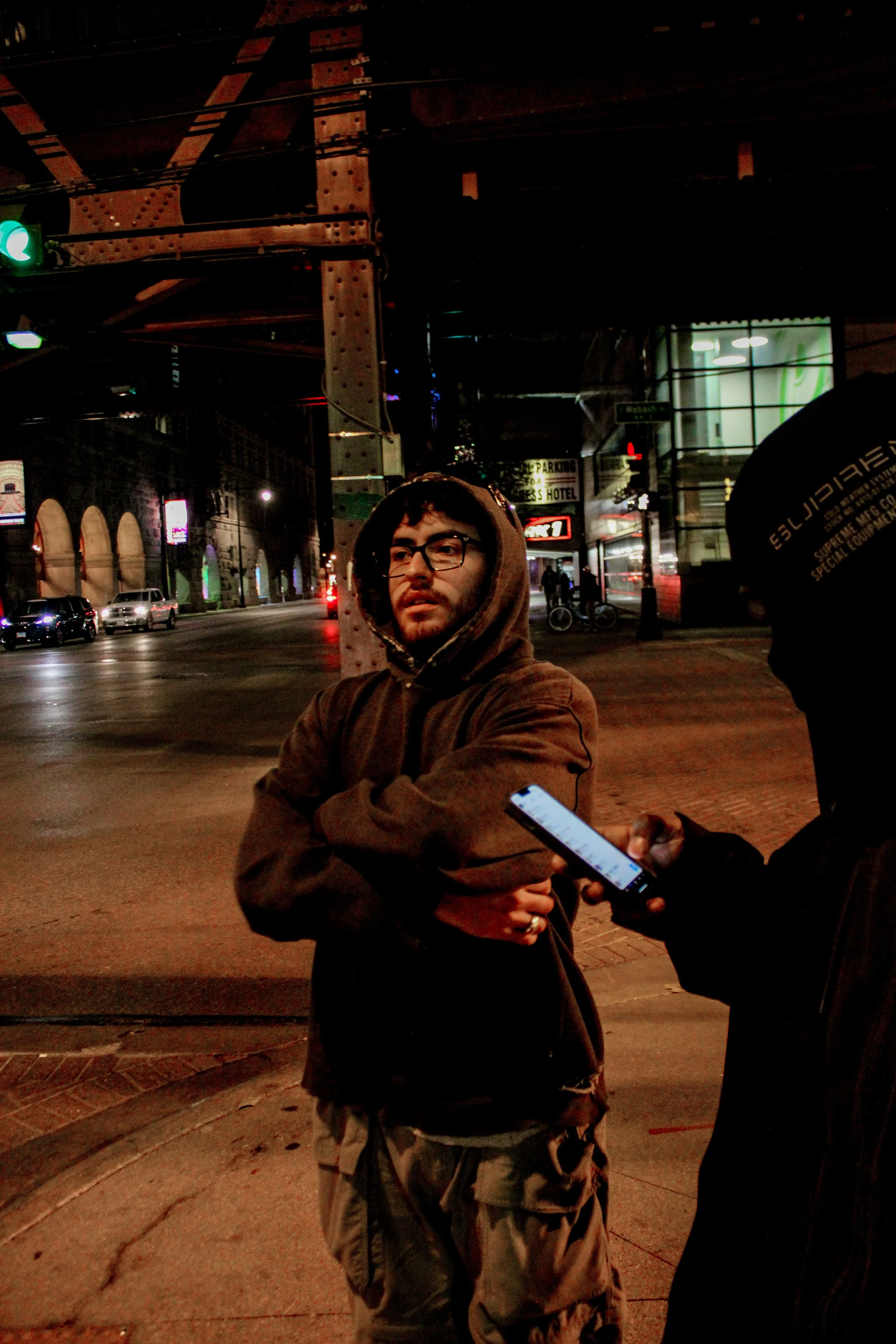 Nighttime street scene with two men, one in a hoodie talking on a phone, the other from behind holding a device, under a bridge or elevated track, with neon signs and cars in the background.