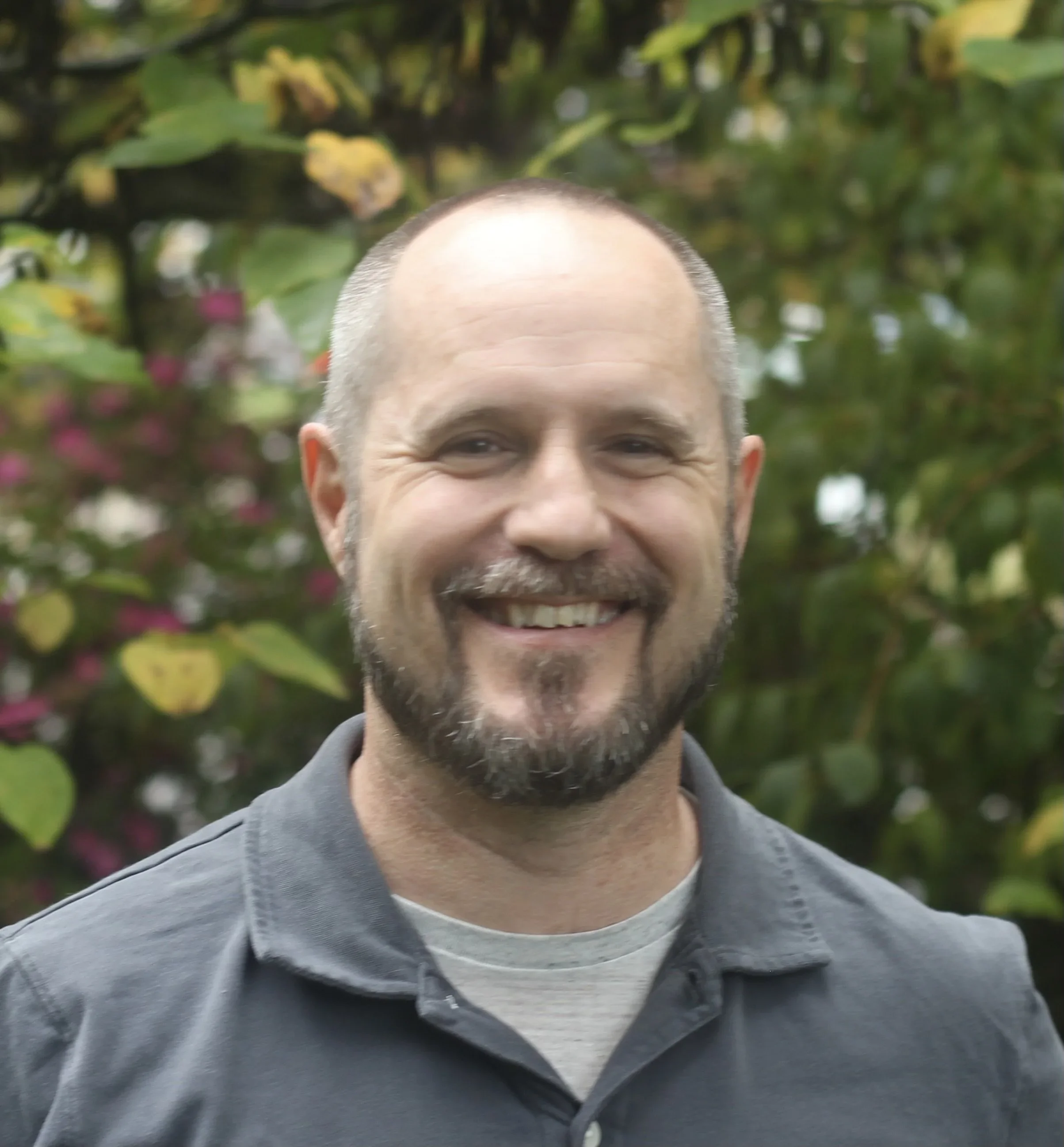 A smiling man with a beard and short hair standing outdoors, with green foliage and pink flowers in the background.
