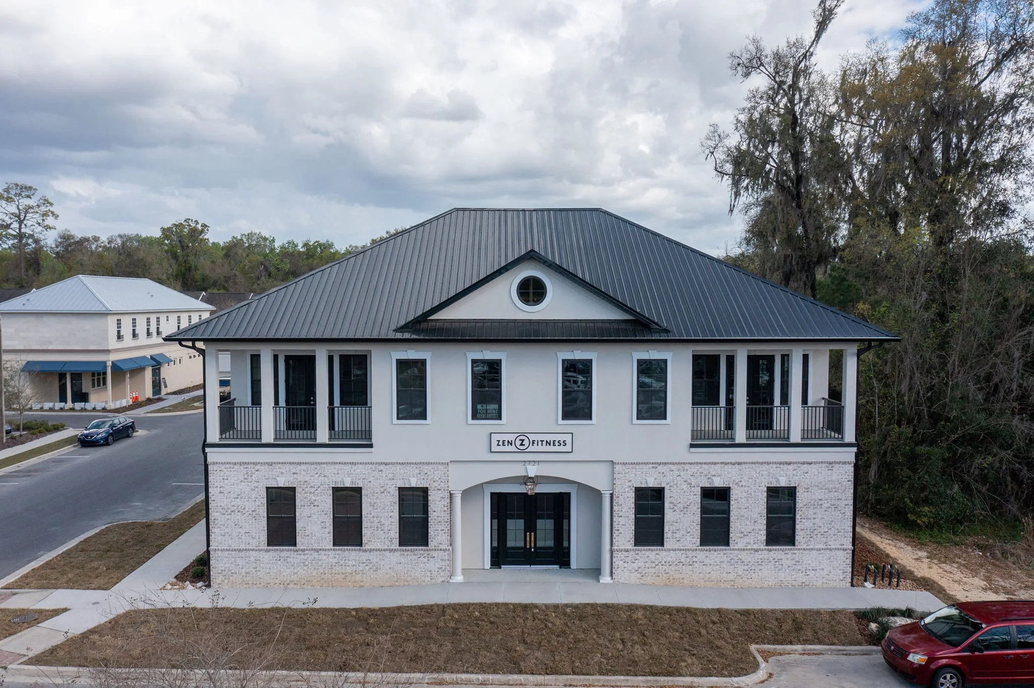 A two-story white building with black window frames and a black metal roof, labeled 'Zen Fitness,' with a small parking area and trees in the background.