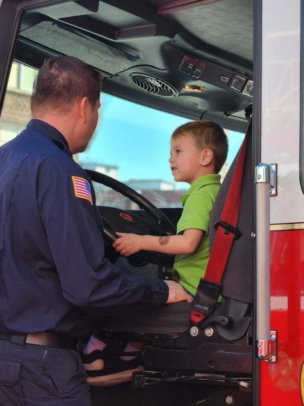 A firefighter with an American flag patch on his uniform kneels next to a young boy sitting in the driver's seat of a fire truck, holding the steering wheel and talking to him.