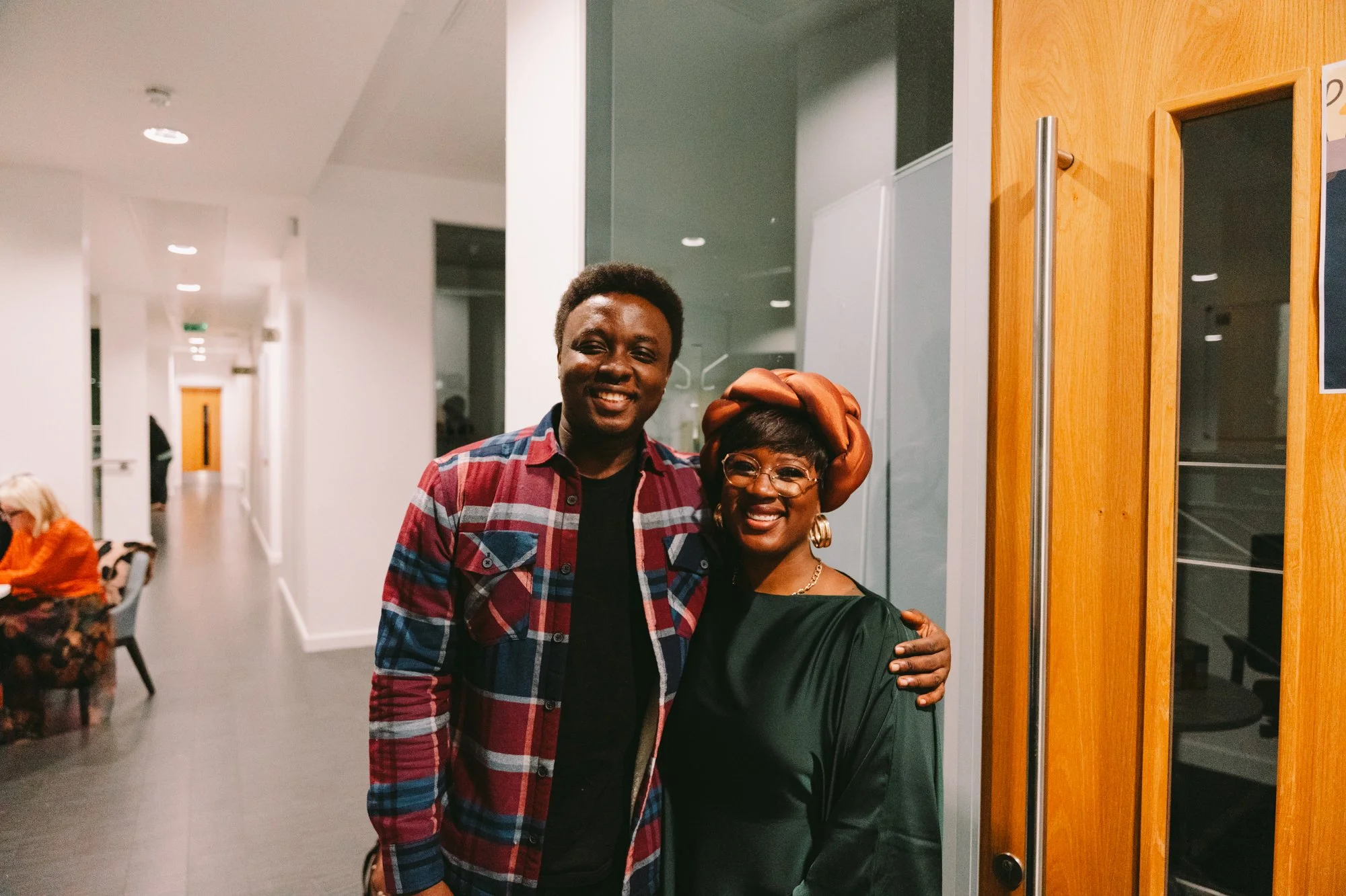 A woman and a man smiling and posing together indoors. The woman is wearing glasses, a large headwrap, and a dark green top. The man is wearing a plaid shirt over a black t-shirt.