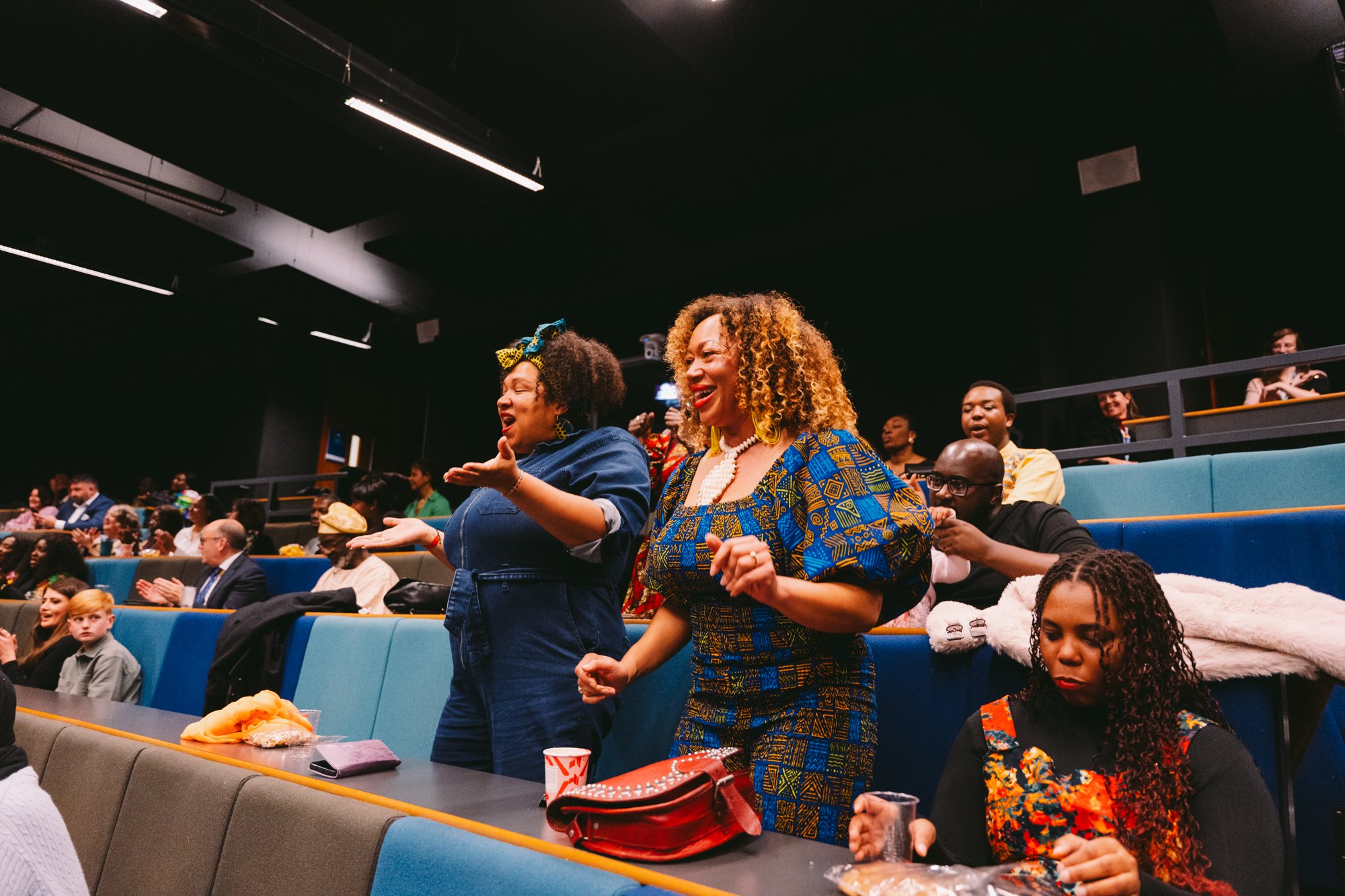 People standing and singing or cheering in an auditorium or theater with people seated in the background.