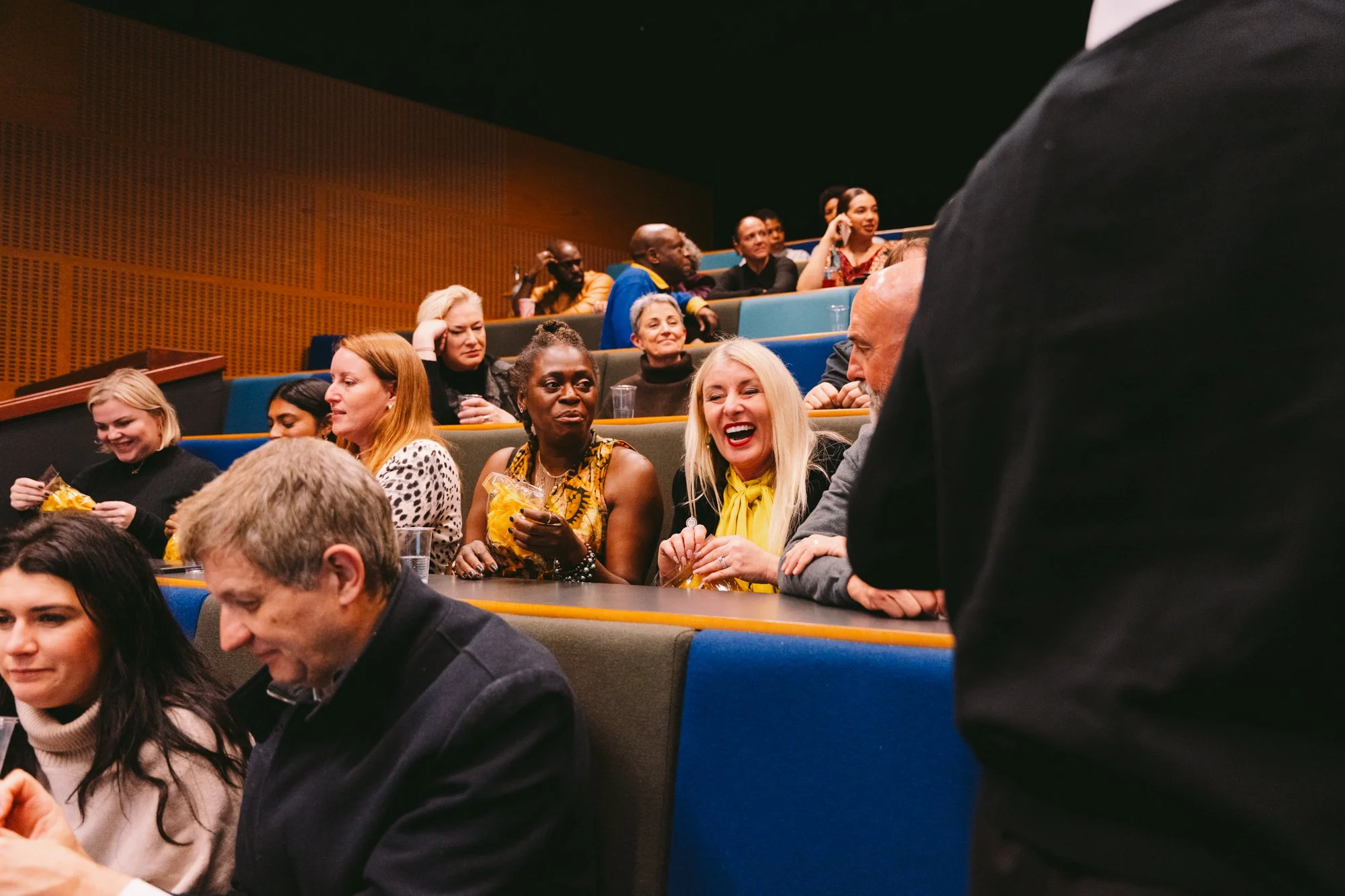 People sitting in an auditorium, some smiling and chatting, with a speaker standing in front of them.