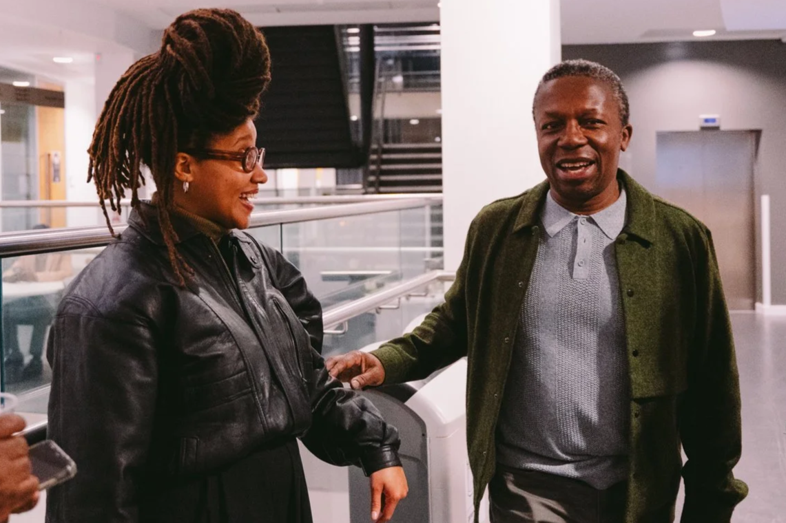 A woman with dreadlocks and glasses smiling and talking with a man in a gray polo shirt and green jacket in a modern indoor setting.