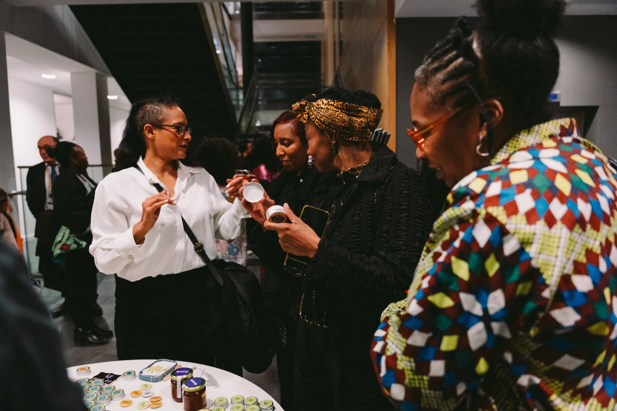 Group of women at a social event, chatting and examining jars of products on a table, indoors in a modern building.