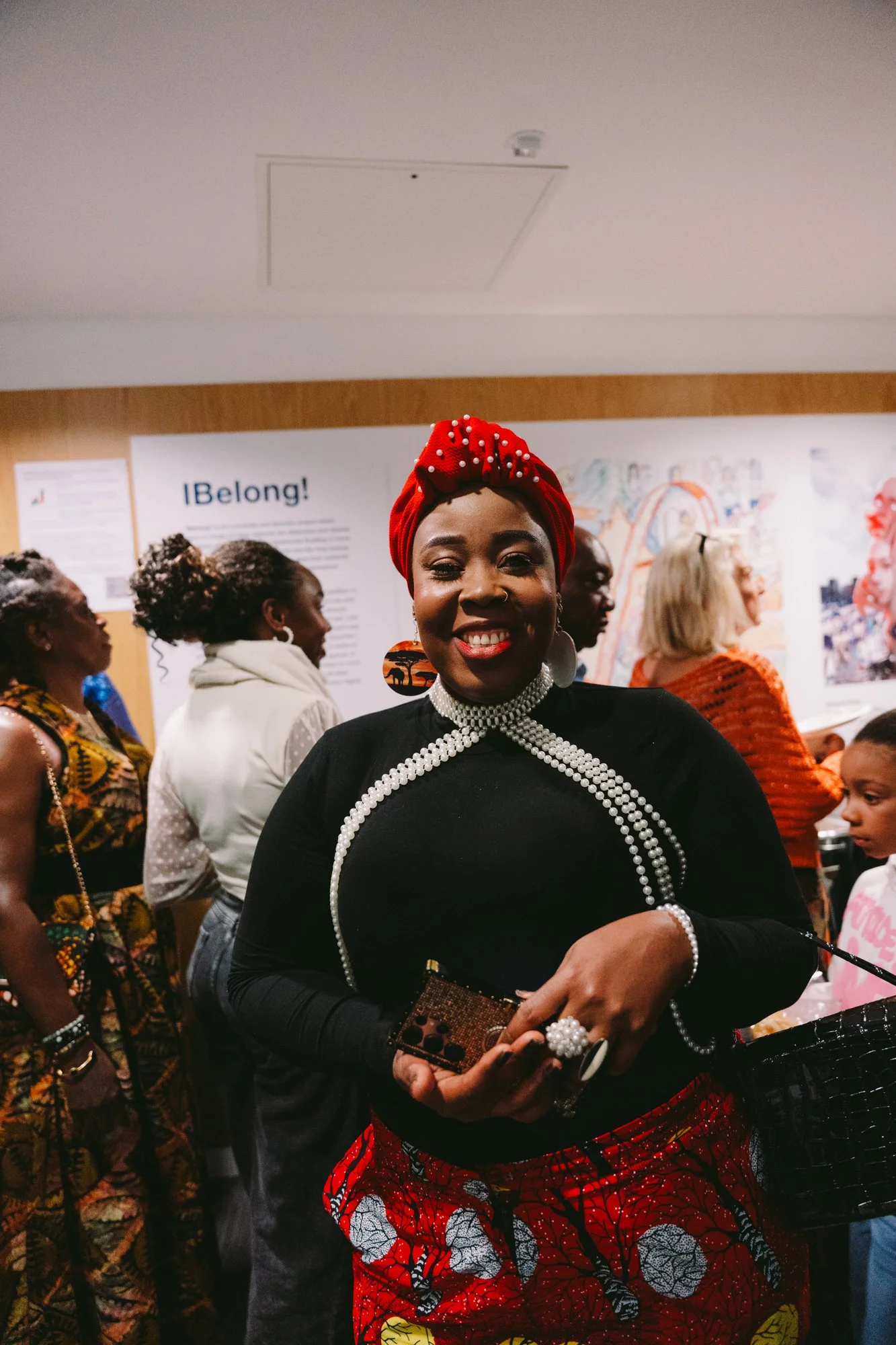 A smiling Black woman wearing a red headwrap, pearl jewelry, and a black top, surrounded by others in a lively indoor setting with artwork on the wall.