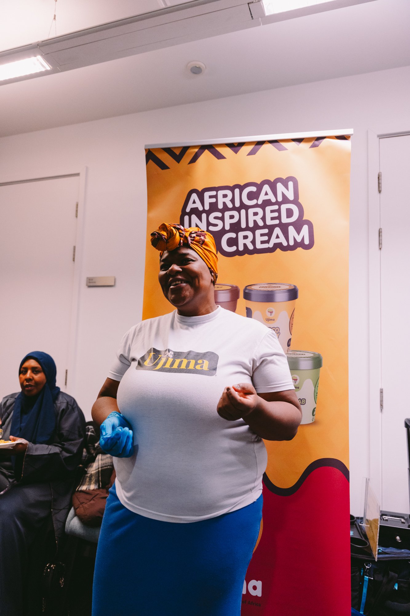 A woman wearing a white T-shirt, a blue skirt, a colorful headwrap, and blue gloves, smiling and posing in front of a yellow and red banner that says "African Inspired Cream" with images of ice cream containers. Another woman in a dark blue hijab is 