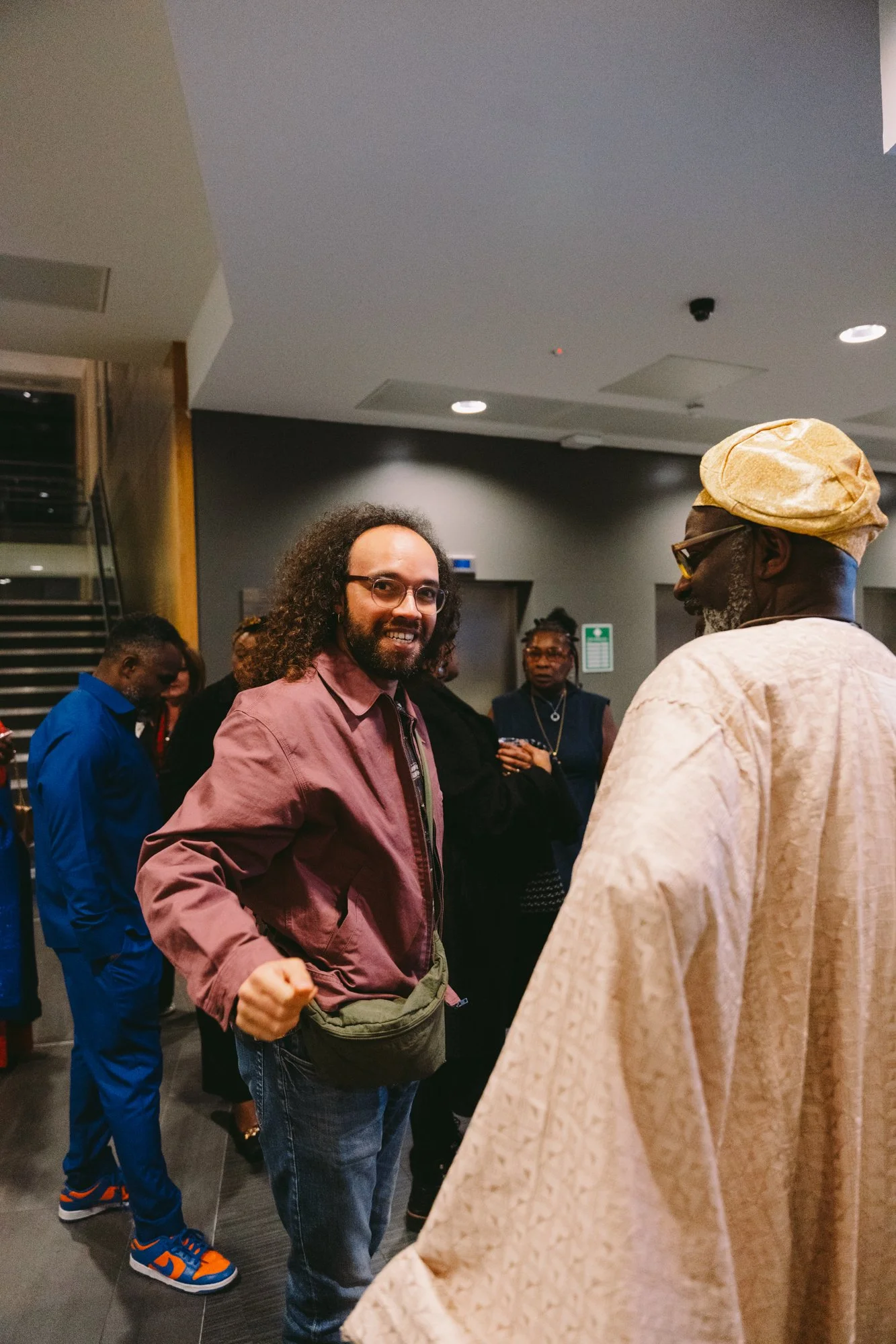 A group of people attending an indoor event, with two men in the foreground engaged in conversation. One man has curly hair, glasses, a maroon jacket, and a crossbody bag. The other man has gray hair, glasses, a beige traditional outfit, and a matchi