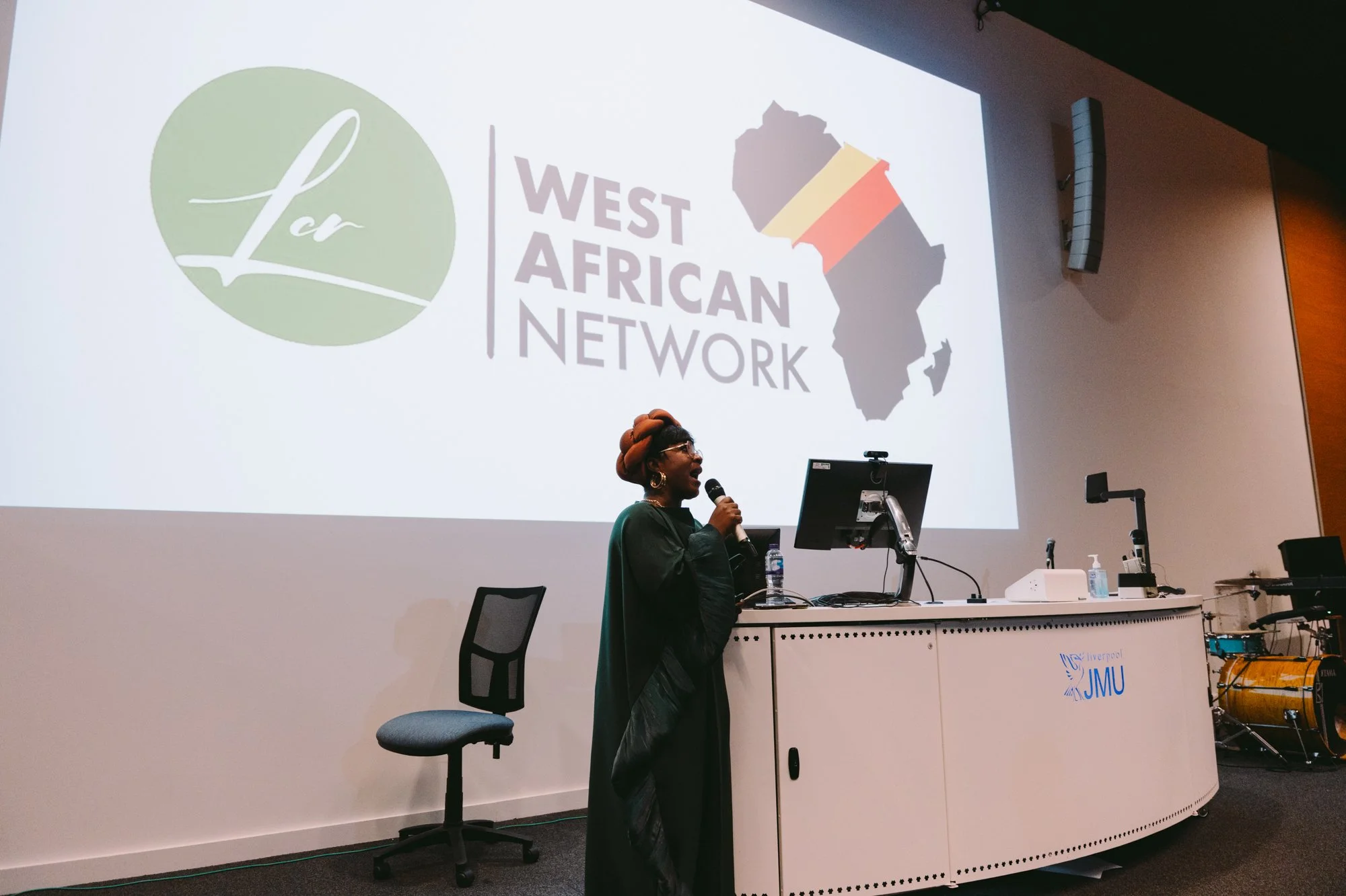 Woman in black traditional attire speaking into a microphone in front of a projected presentation slide with logos for Liverpool Le and the West African Network, and a map of Africa with the Congolese flag colors.