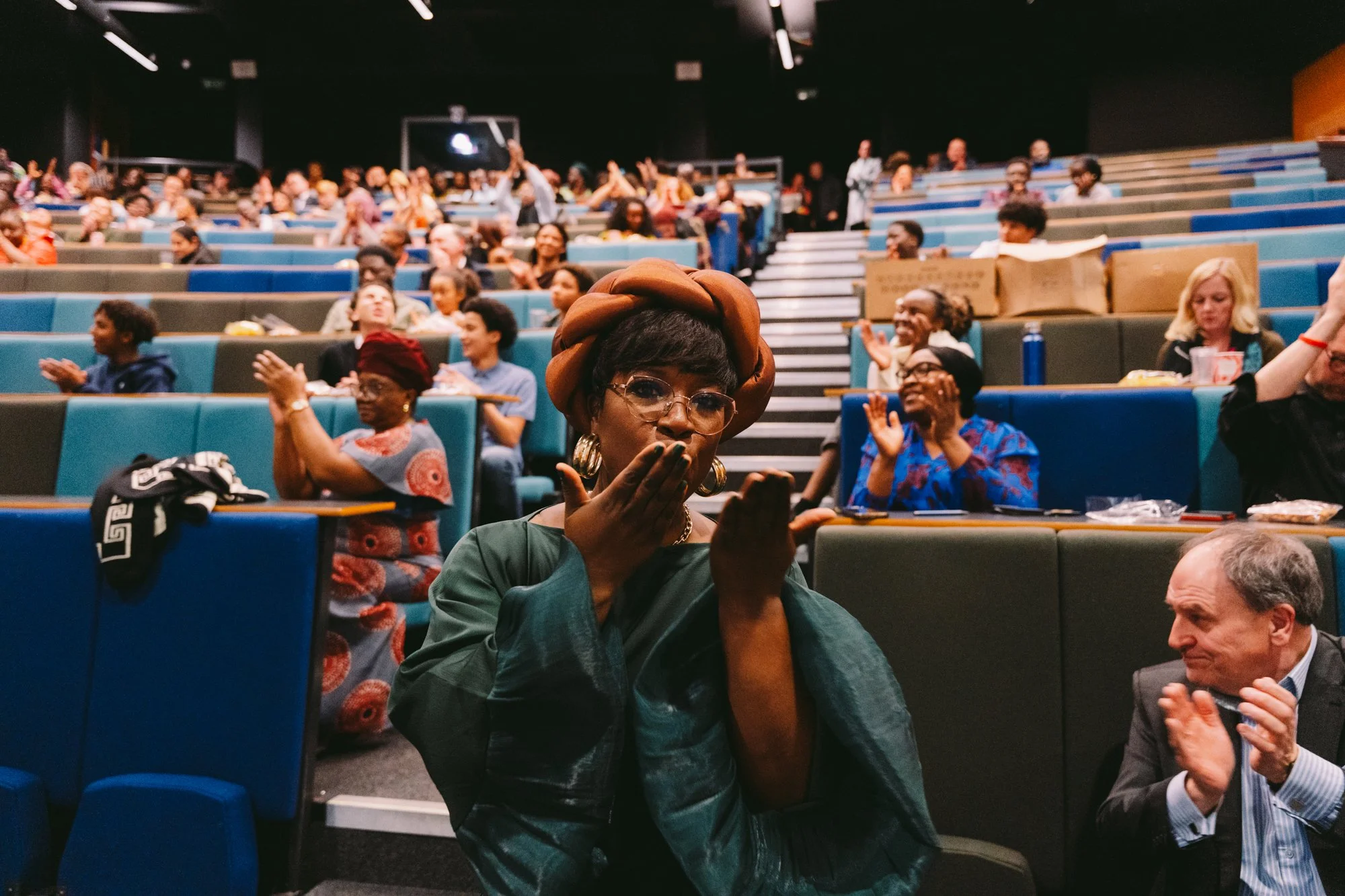 A woman in traditional African attire with a braided headwrap blowing a kiss in an auditorium filled with diverse seated attendees clapping and smiling.