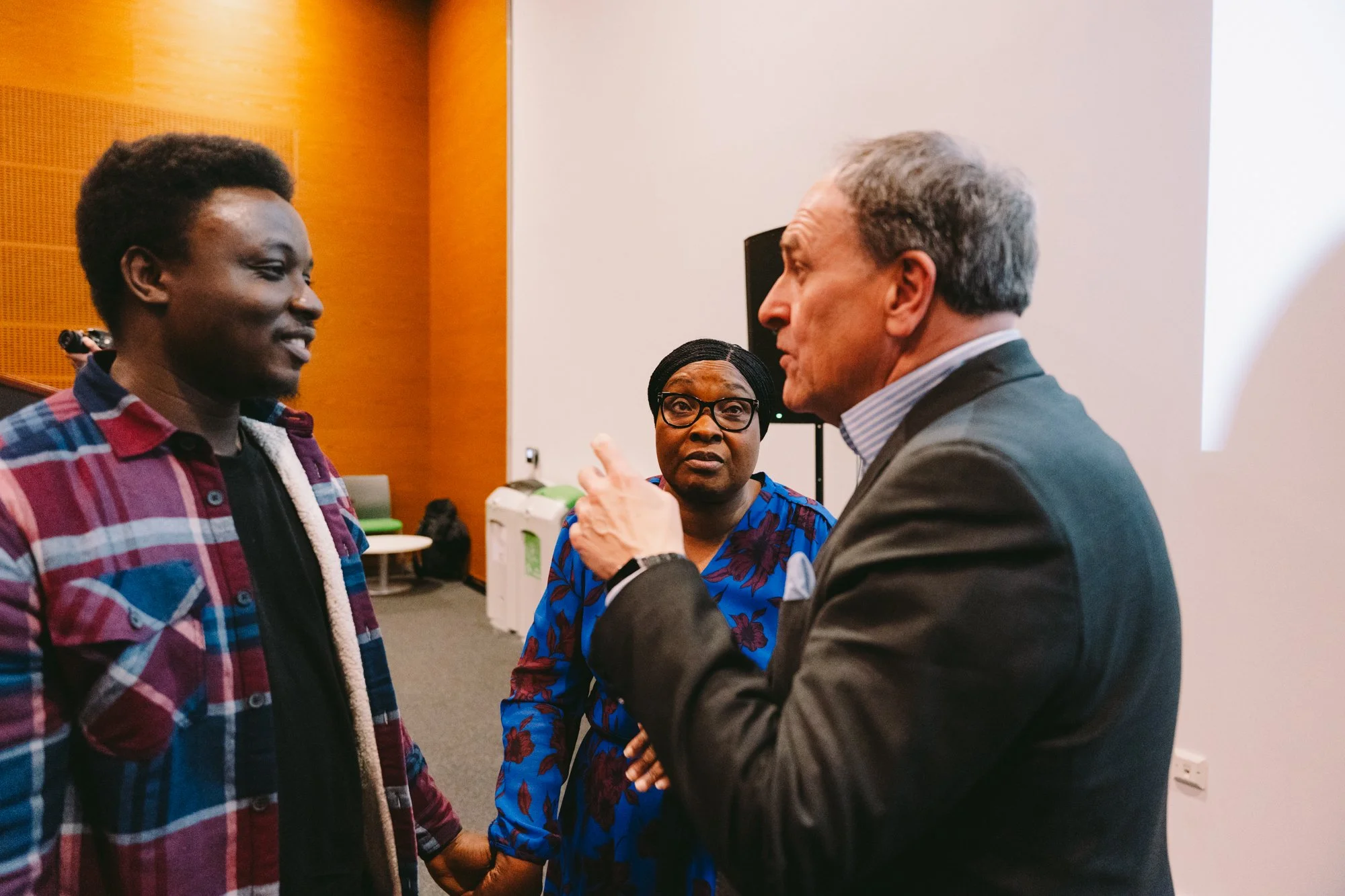 A group of three people engaged in conversation in a room. The man on the right is speaking, with a serious expression. The woman in the middle looks surprised or concerned, and the young man on the left is smiling and listening.